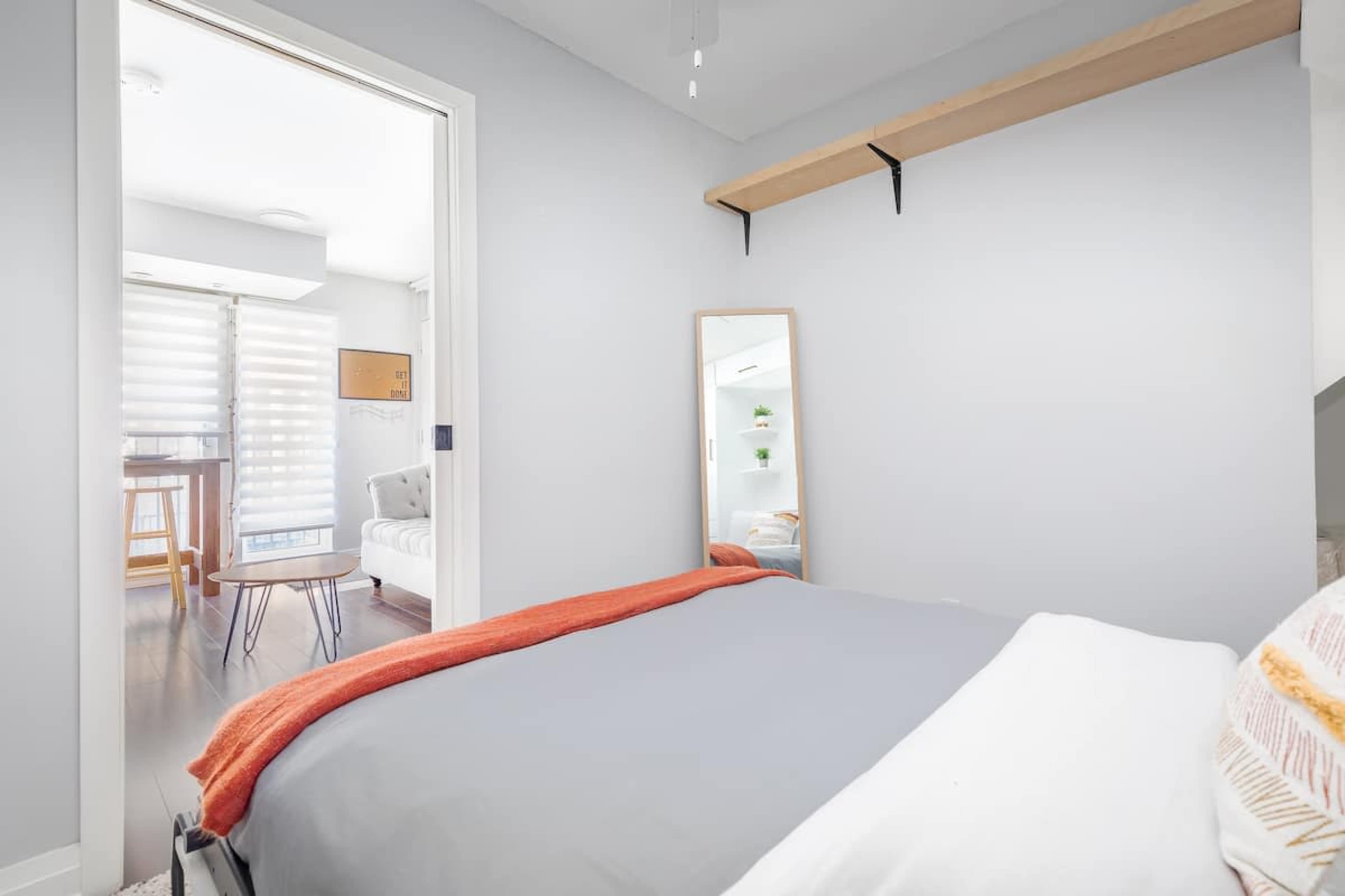 A bedroom with a gray bedspread, a mirror on the wall, and a small table near an open doorway leading to a living area with a sofa.