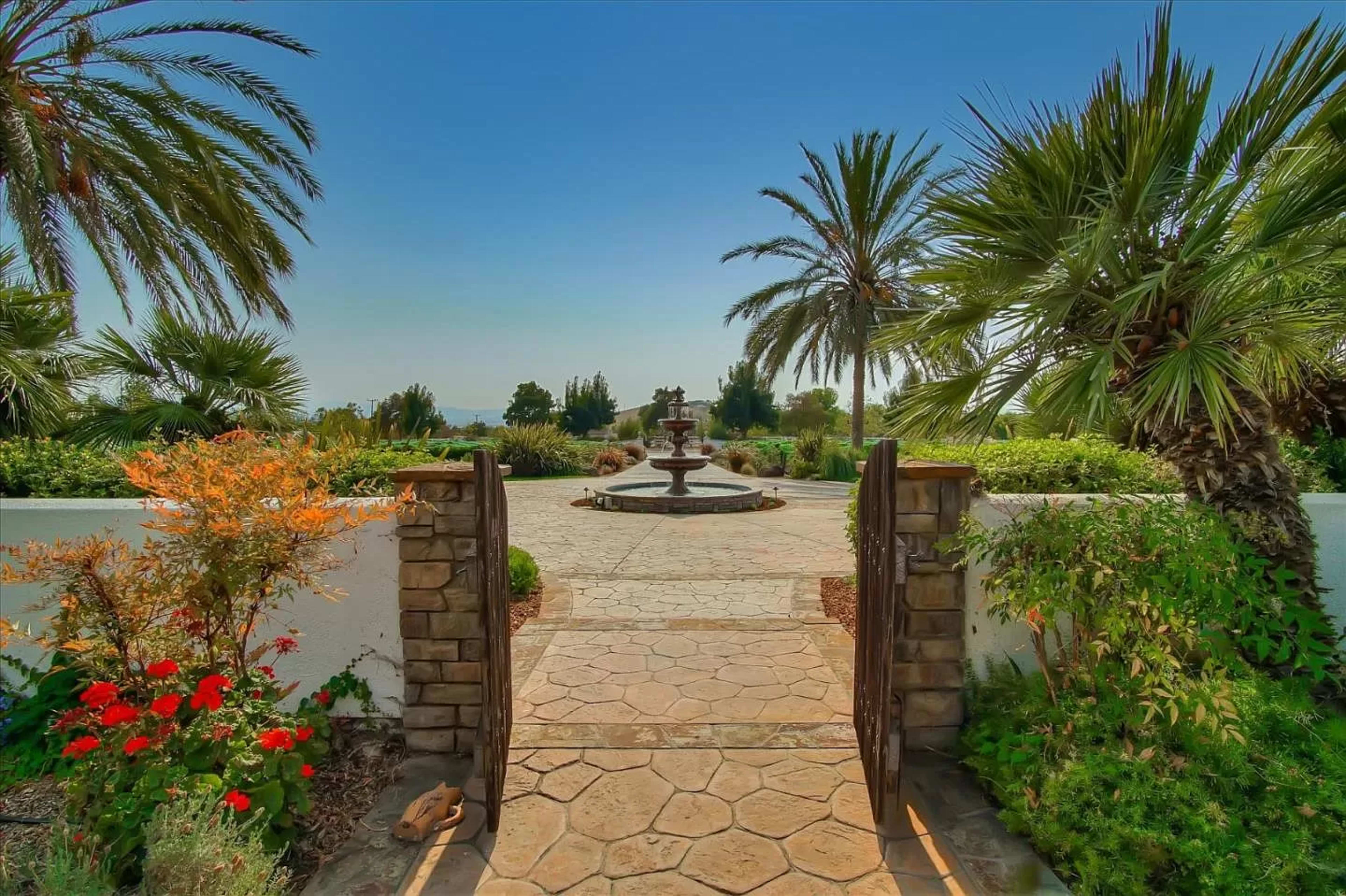 A stone pathway leads through a garden gate to a landscaped area featuring a fountain and palm trees.