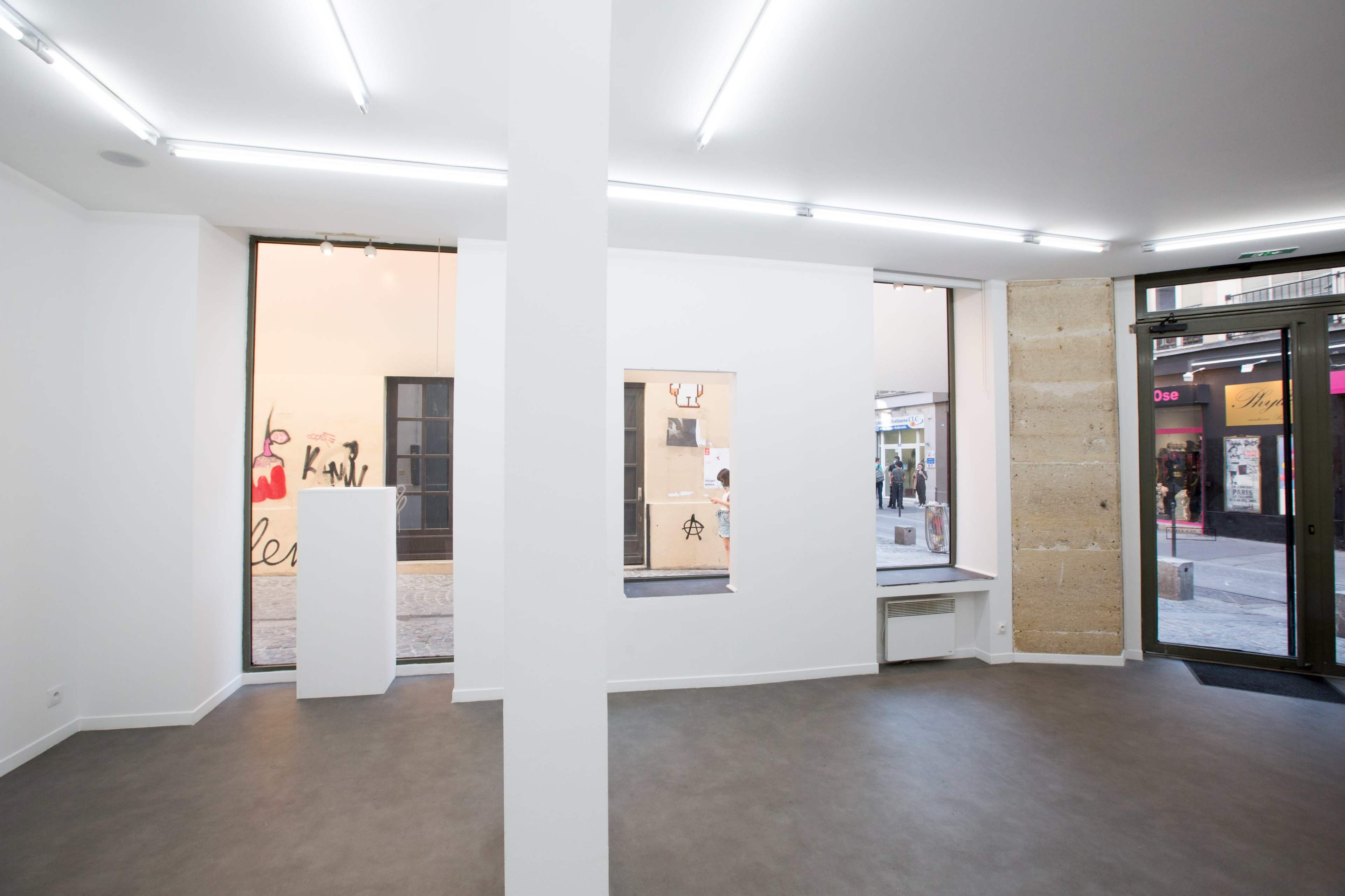The image shows a minimalist gallery space with white walls and floor-to-ceiling windows displaying a street view.