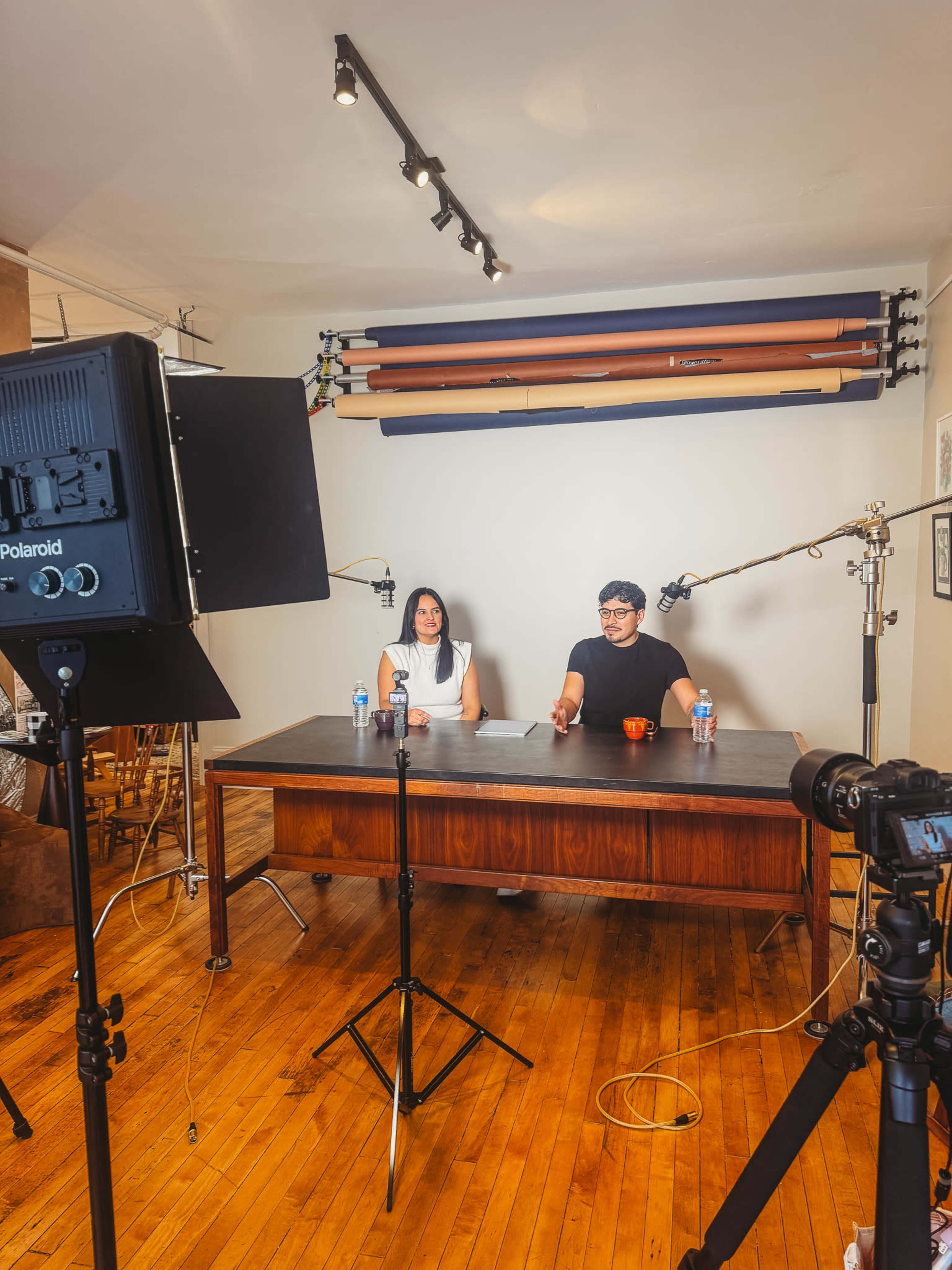 A wooden desk is set up in a room with two people seated behind it, flanked by cameras and studio lights.