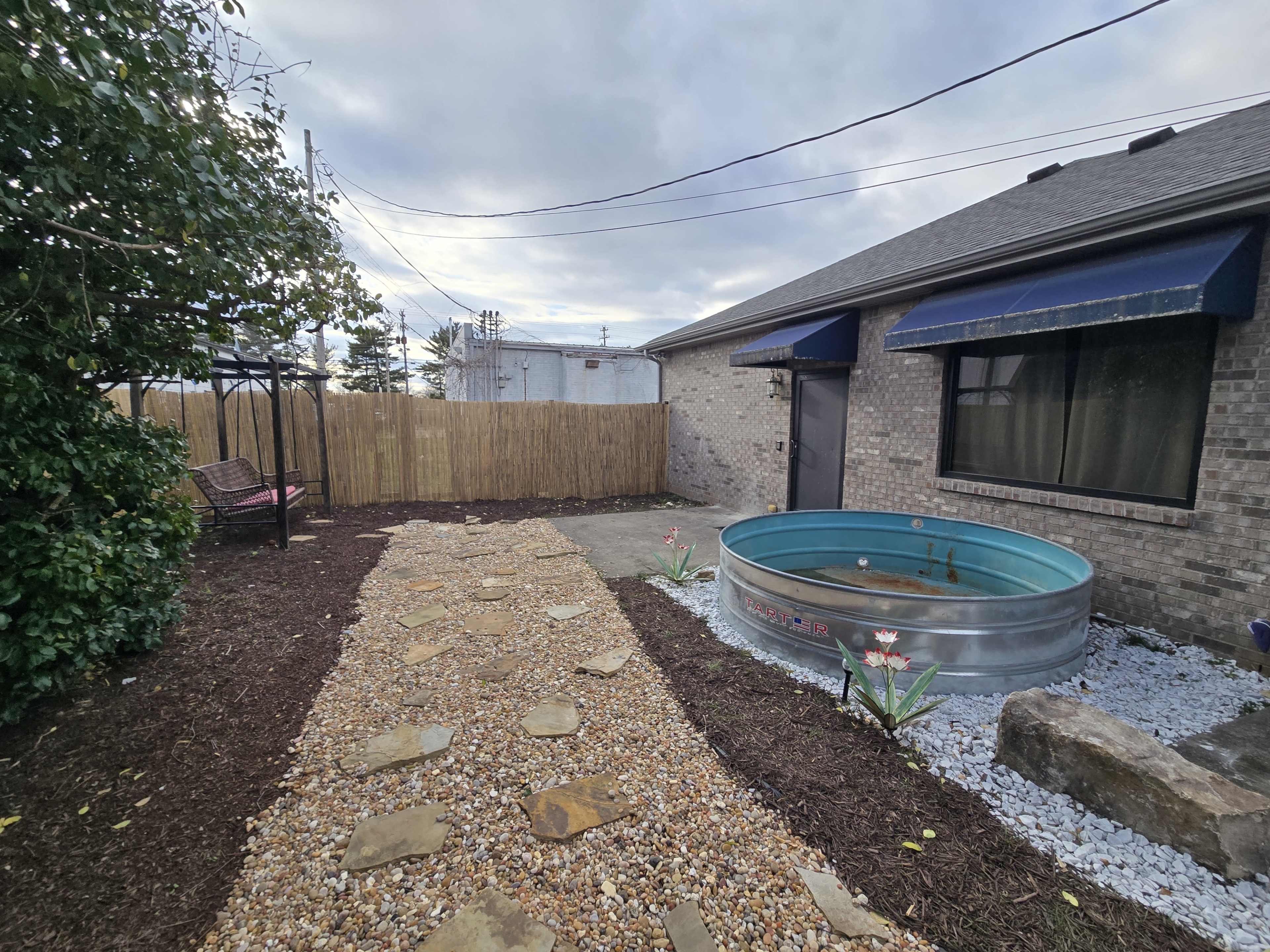 The image shows a backyard pathway made of stones leading to a metal water tank beside a house with a wooden fence and a small seating area.
