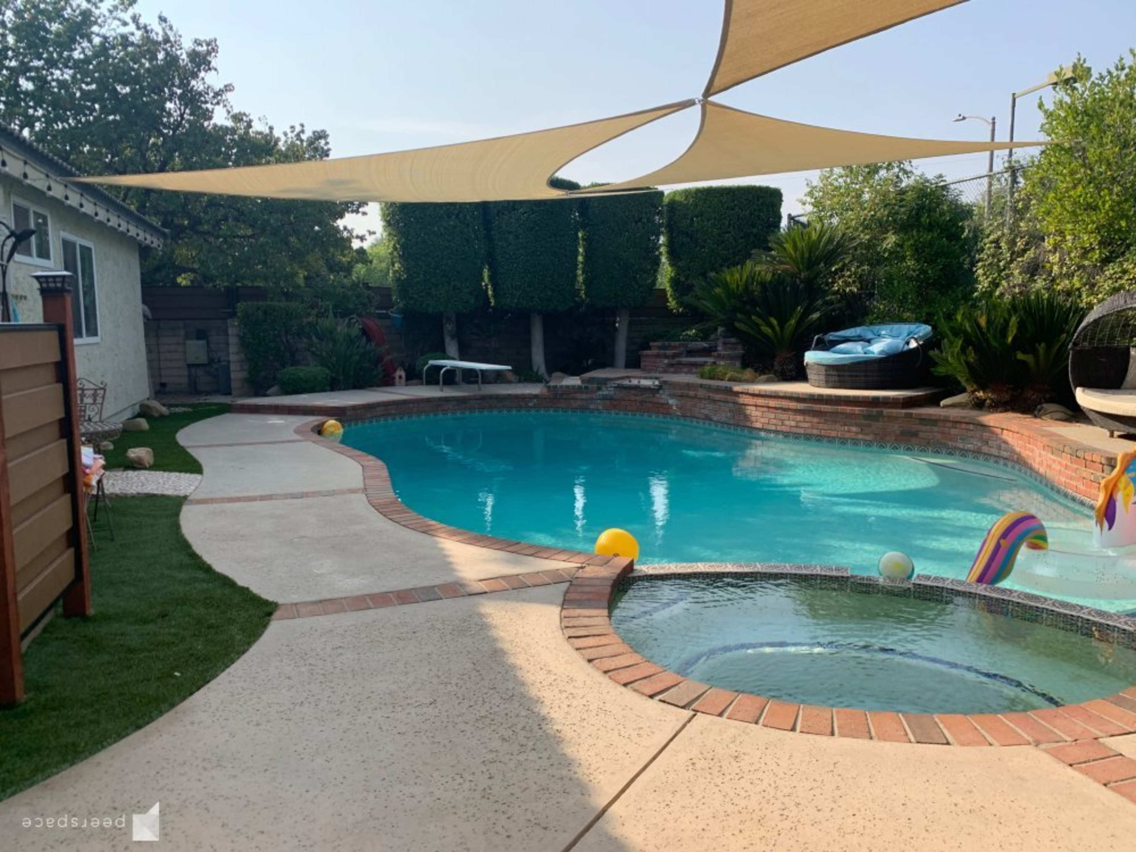 A spacious backyard features a swimming pool surrounded by a patio, grass, and shaded areas, with colorful pool toys floating in the water.