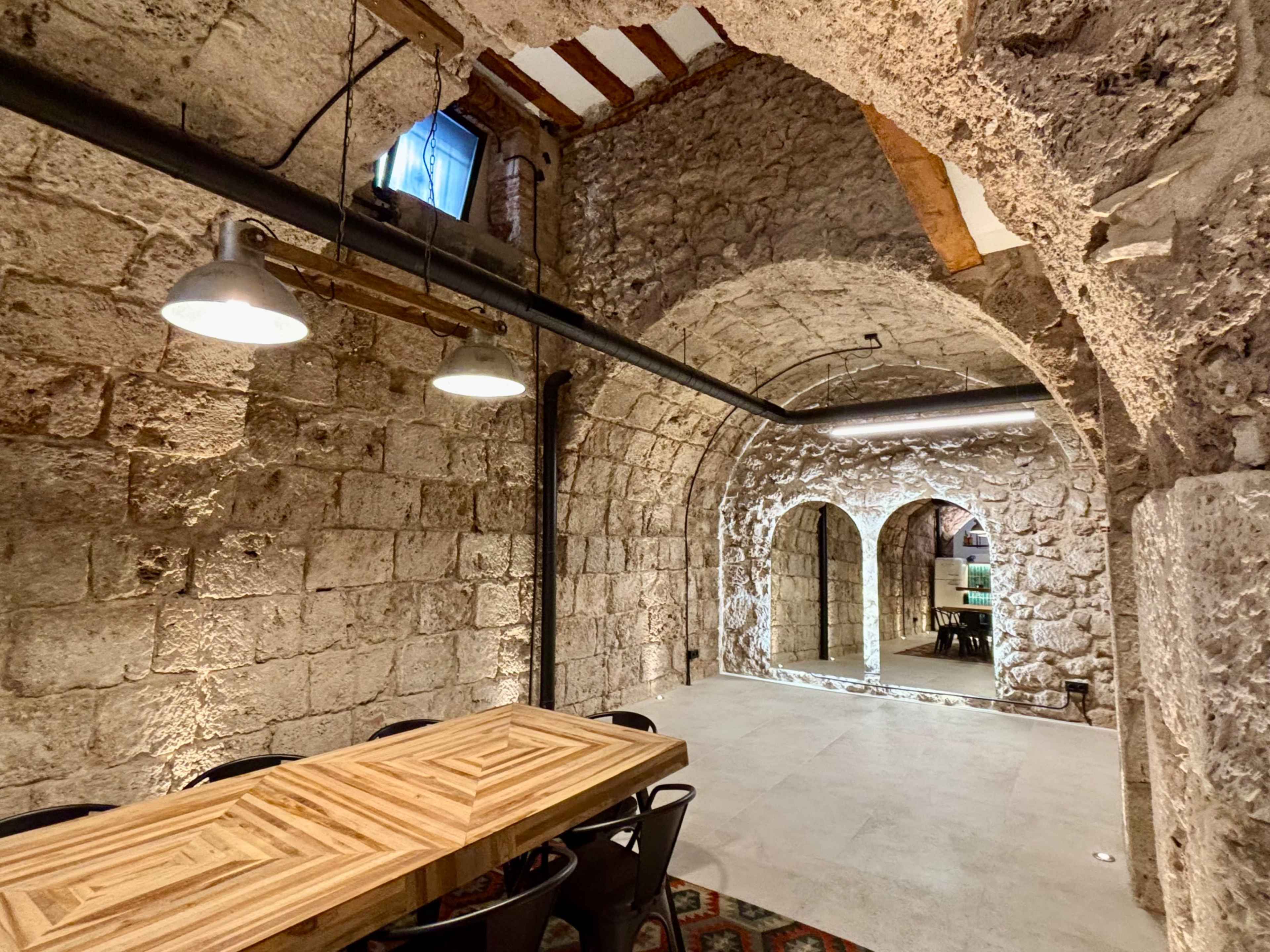 The image features a rustic stone interior with arched ceilings, illuminated by overhead lights, and a wooden table surrounded by black chairs.