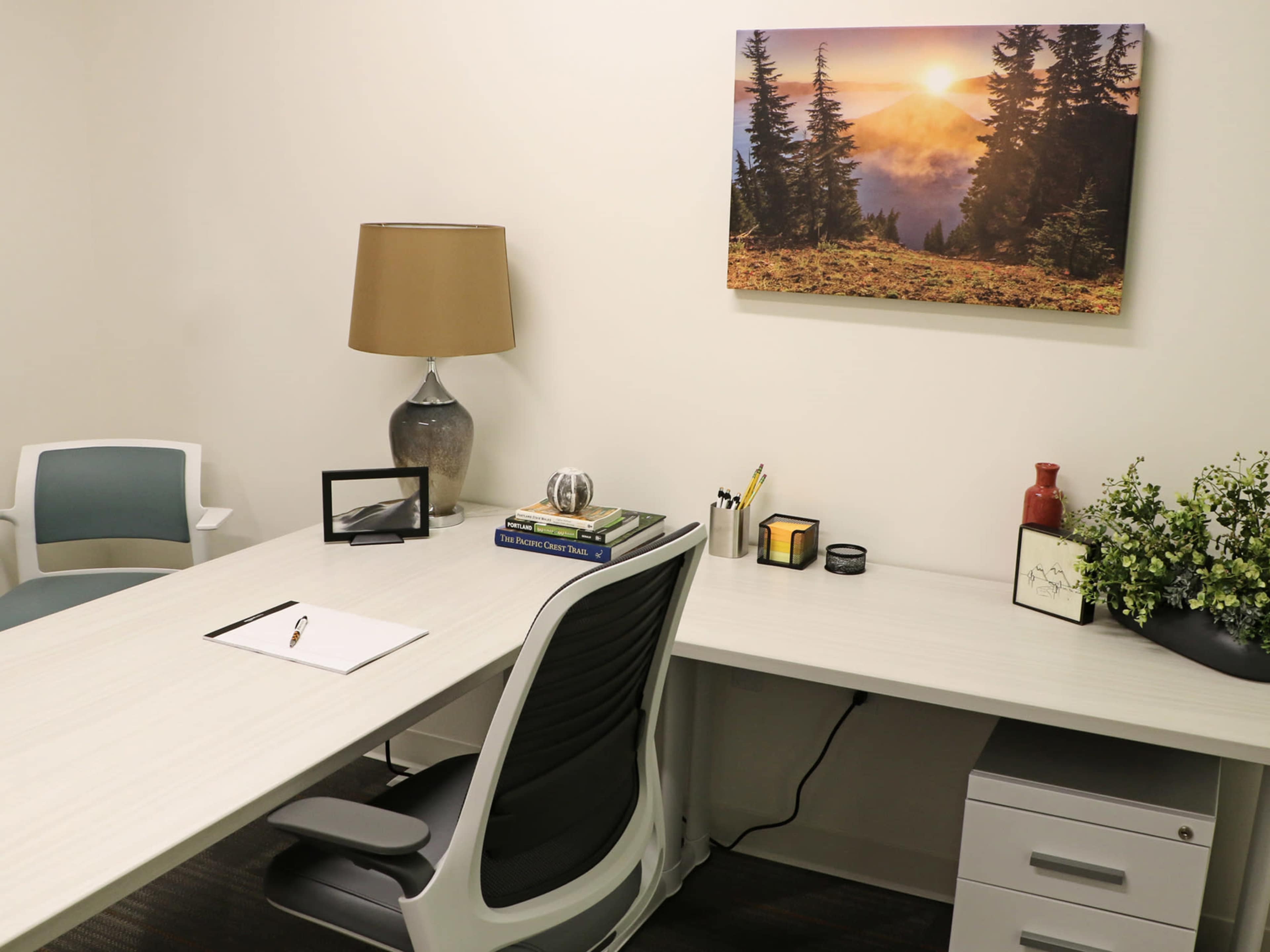 A minimalistic office space features a wooden table with a notebook, a lamp, and decor items, alongside a comfortable chair and desk with a plant and artwork on the wall.