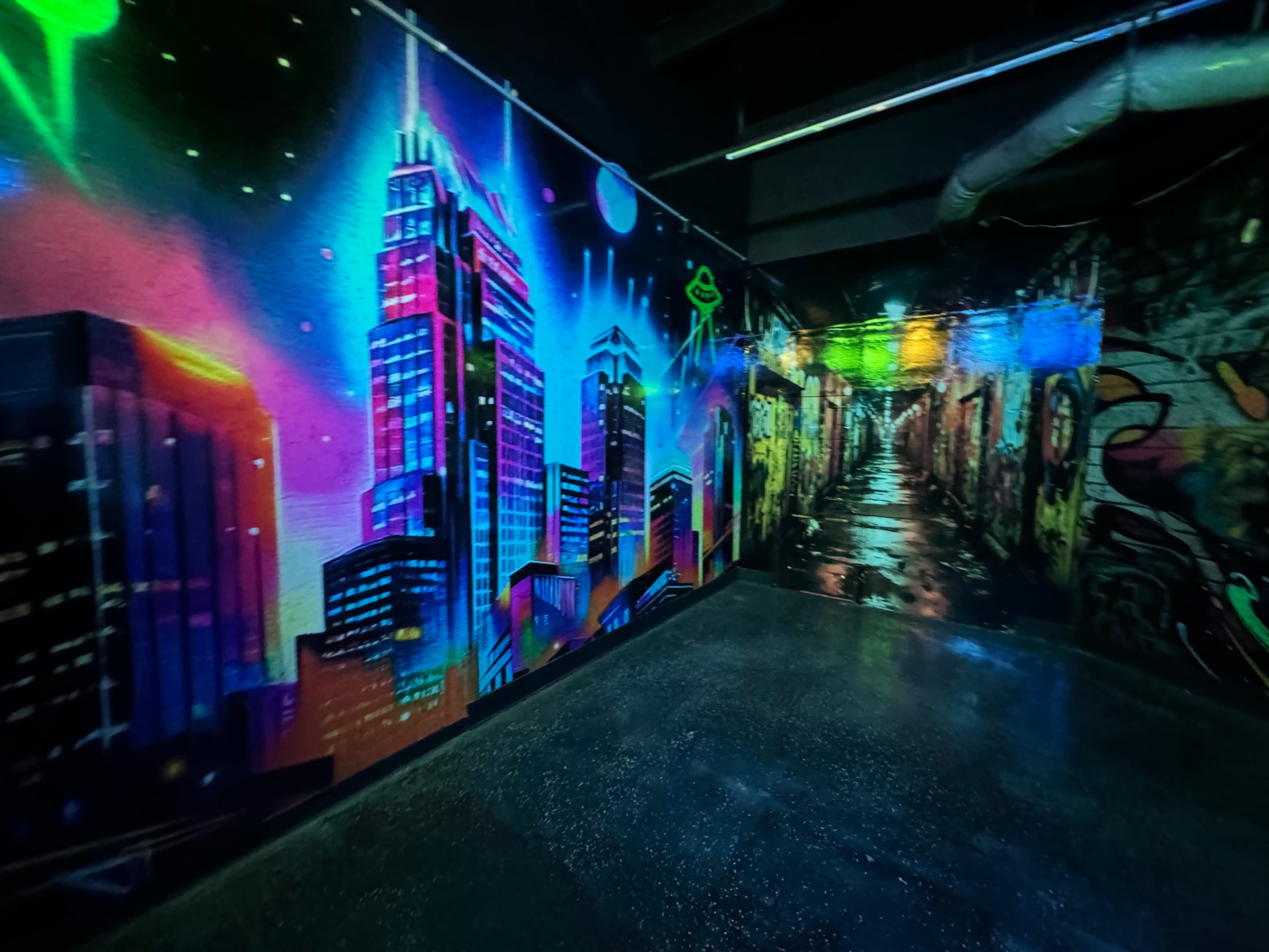 The image shows a brightly colored mural depicting a city skyline on one wall and a dark alleyway scene on the opposite wall, illuminated by vibrant neon lights.
