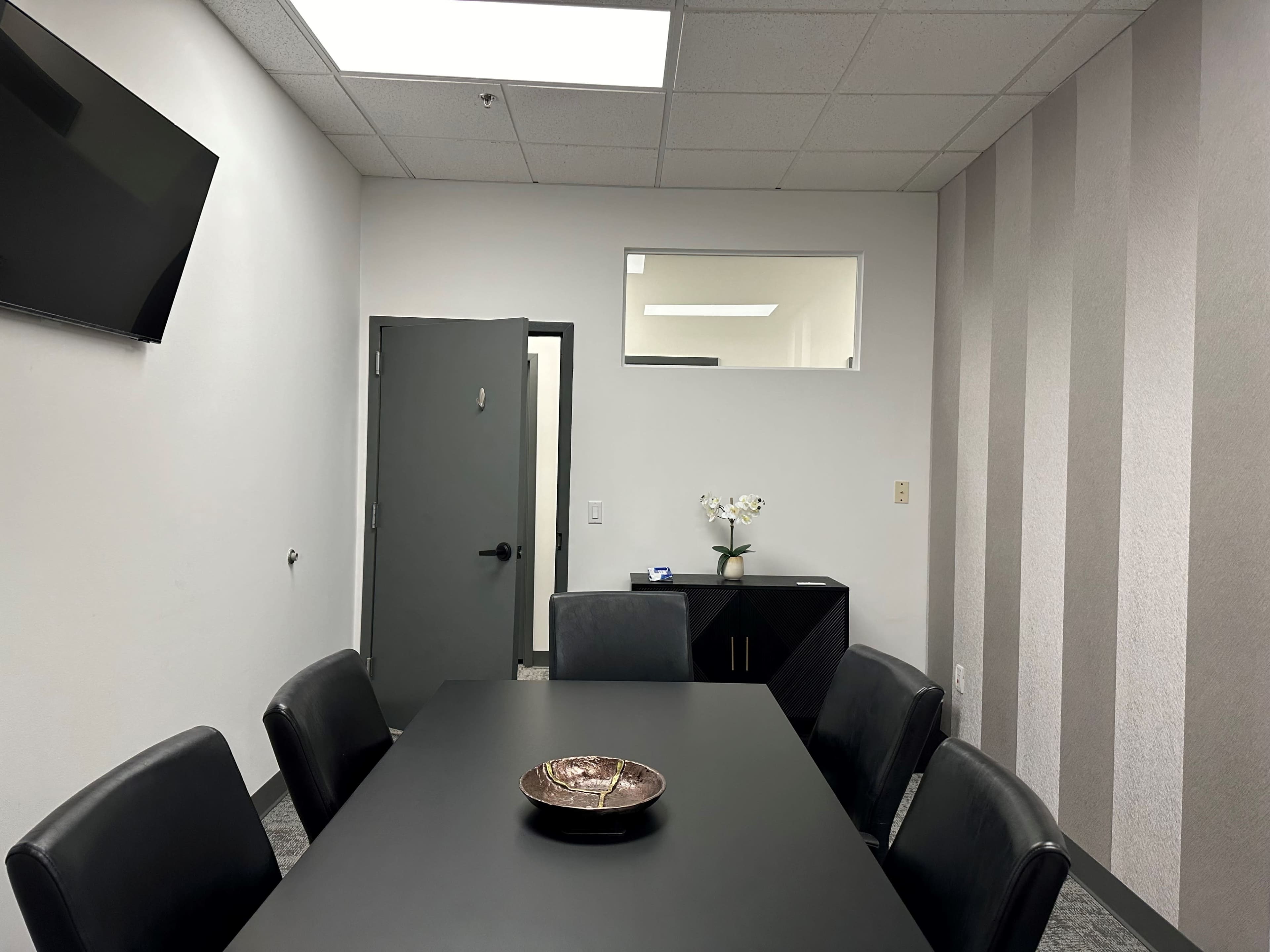 The image depicts a conference room featuring a rectangular black table surrounded by leather chairs, a television mounted on the wall, and a cabinet with a small vase of flowers.
