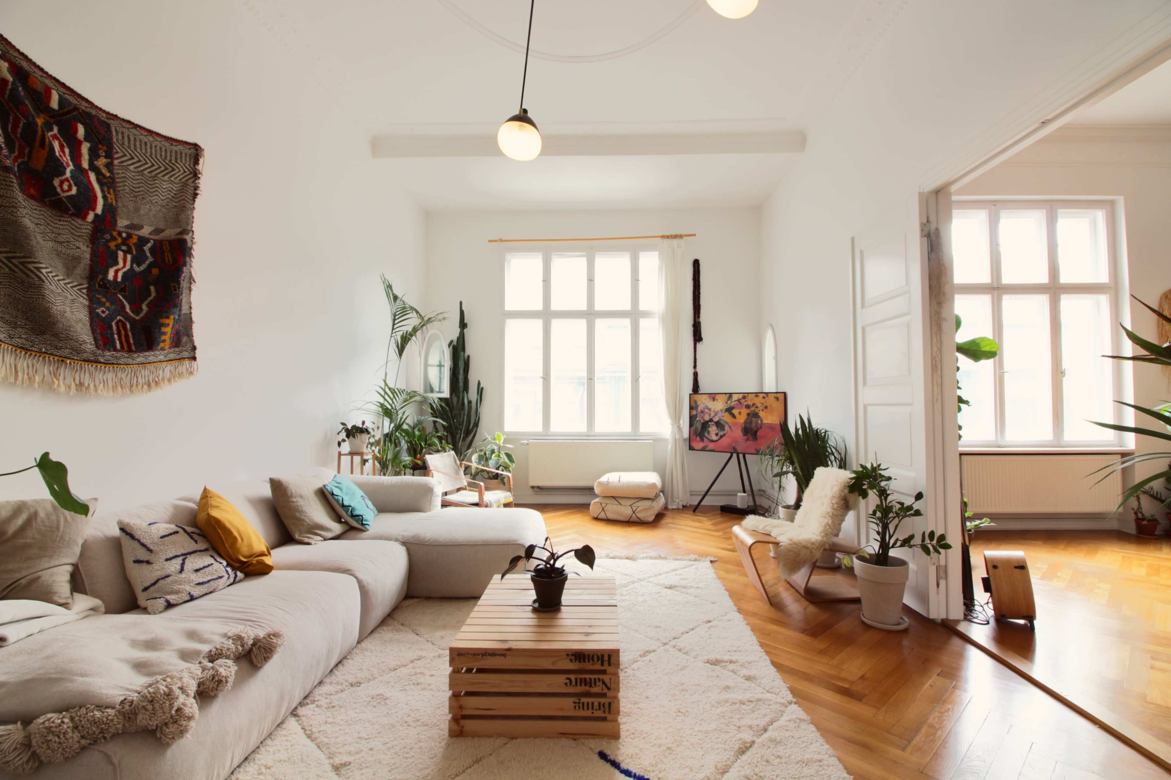 The image shows a bright, minimalistic living room with a large sofa, a wooden coffee table, and several indoor plants.