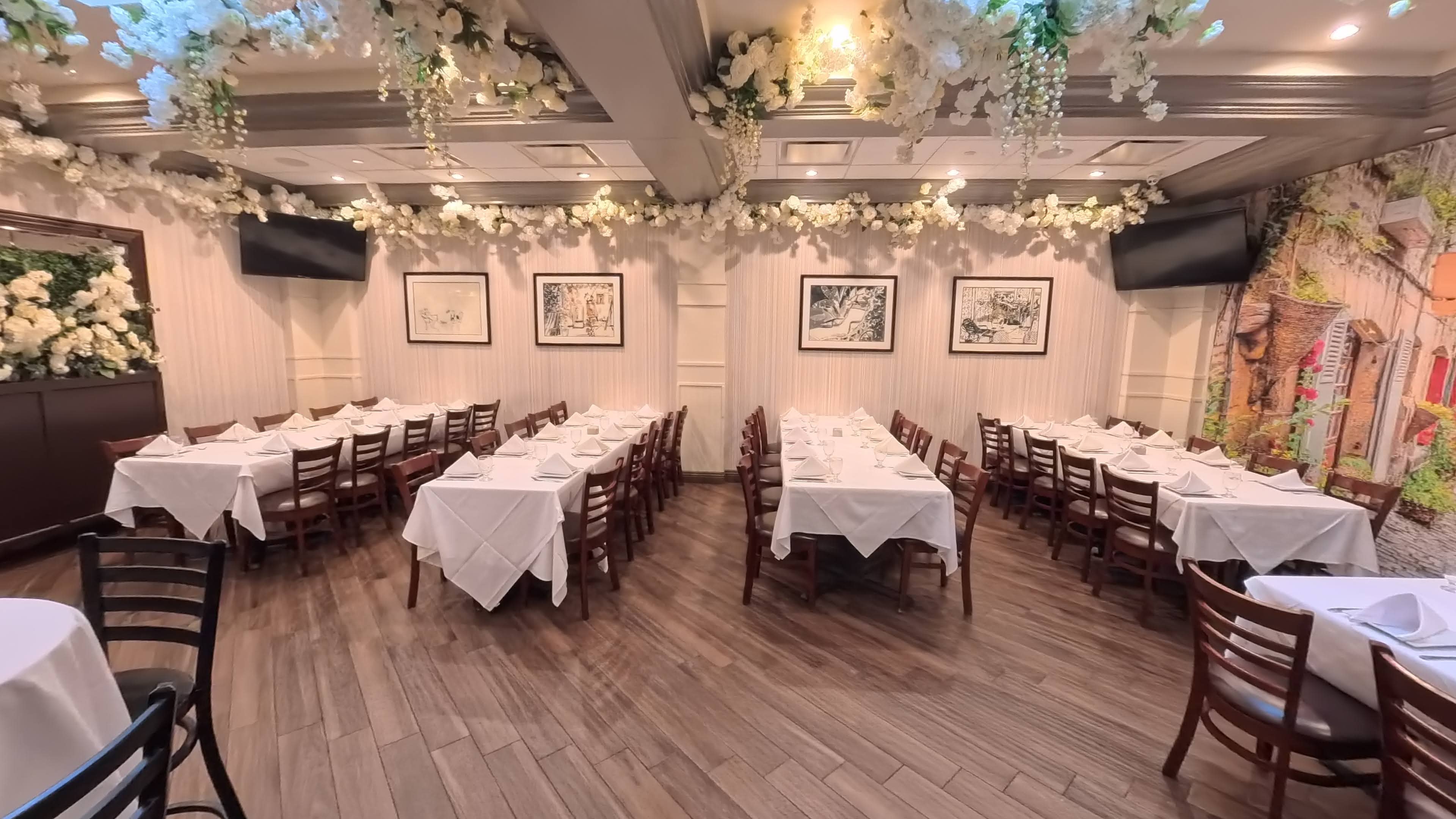 The image shows a dining room in a restaurant with neatly arranged tables, each covered with white tablecloths and surrounded by wooden chairs, against a backdrop of floral decorations on the ceiling and walls.