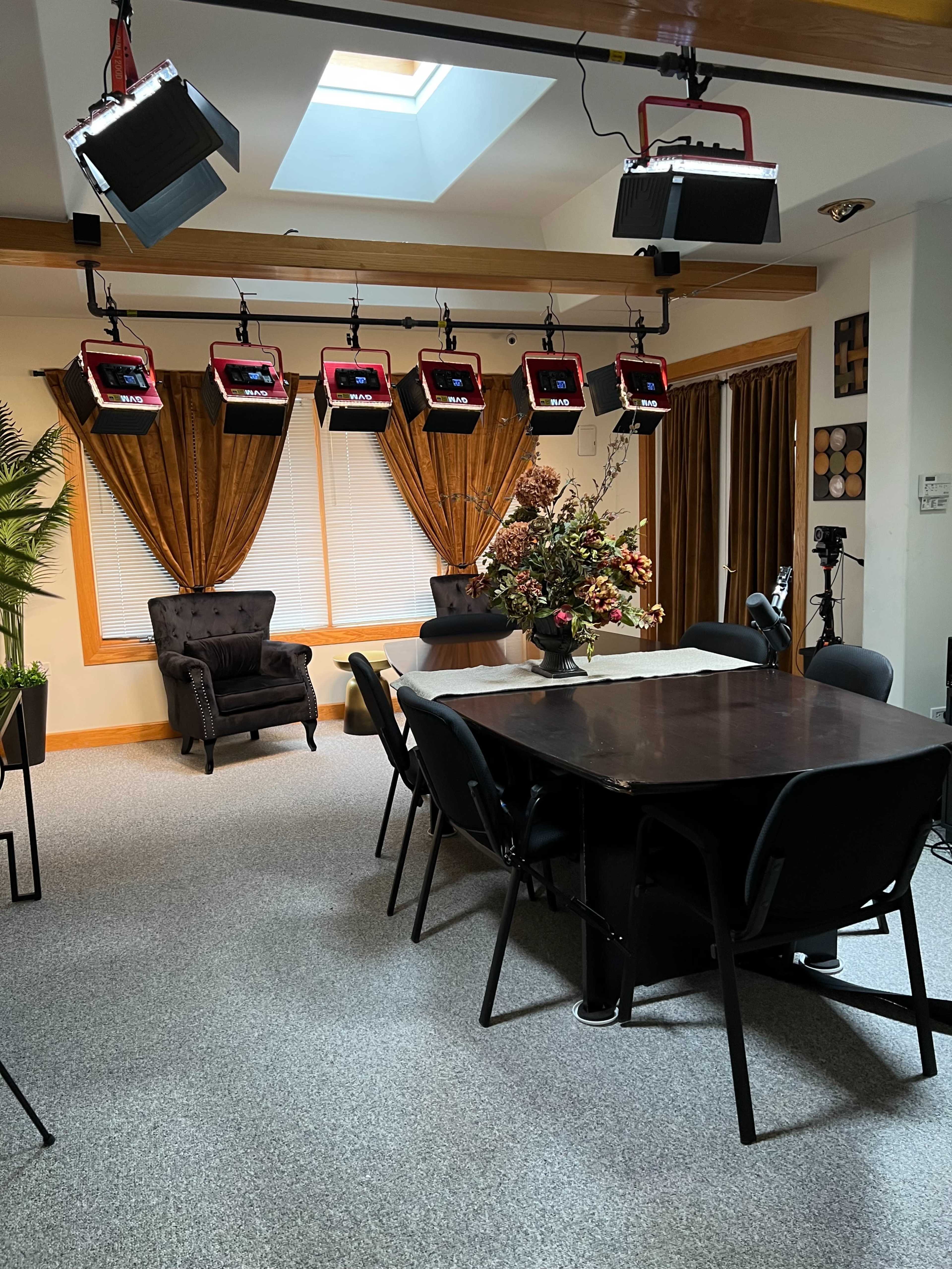 A room with a dining table surrounded by black chairs, focused lighting fixtures overhead, and curtains framing the windows.