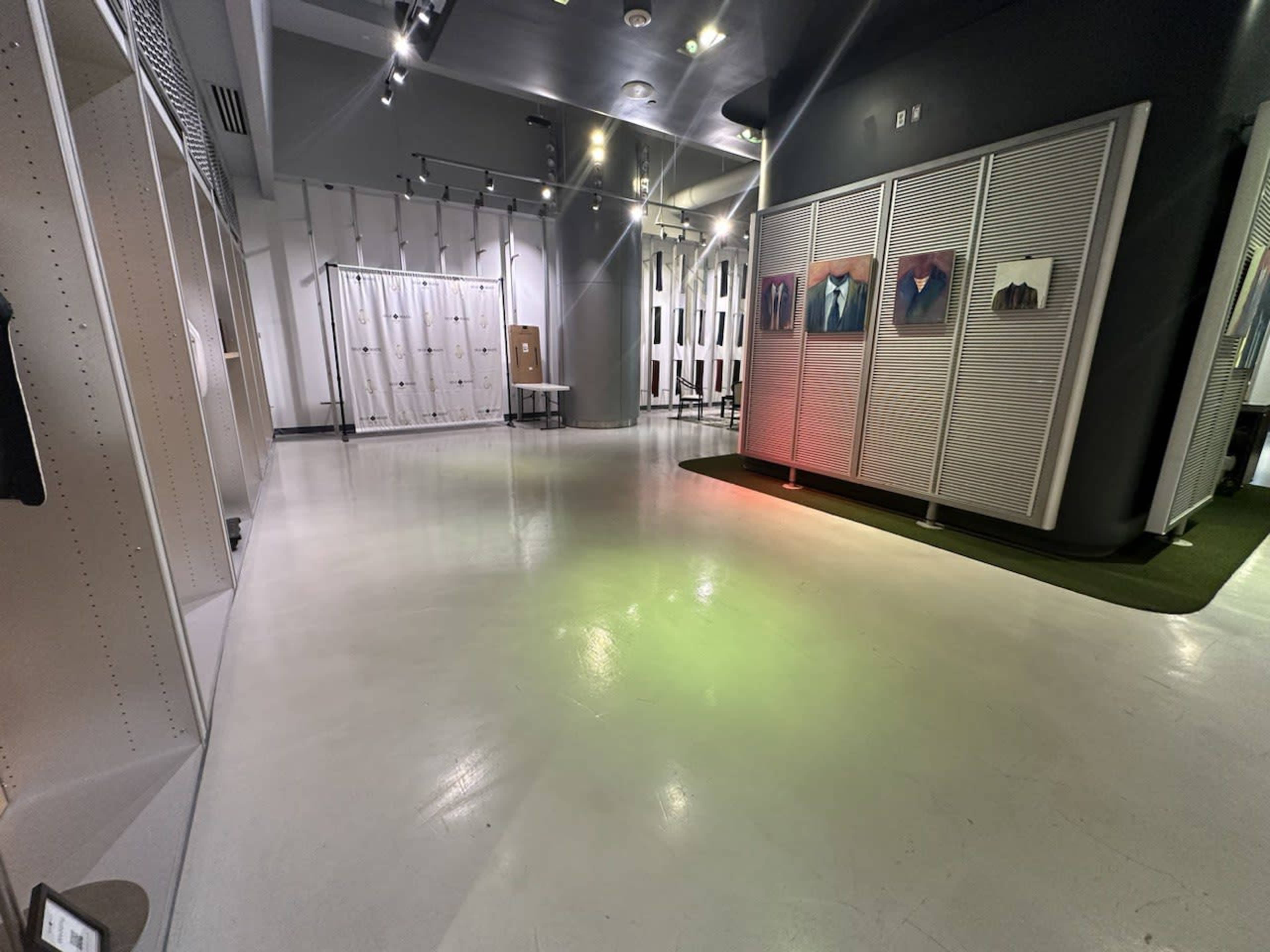 The image shows a spacious, modern exhibition room with white walls, polished flooring, and several display panels featuring artwork.