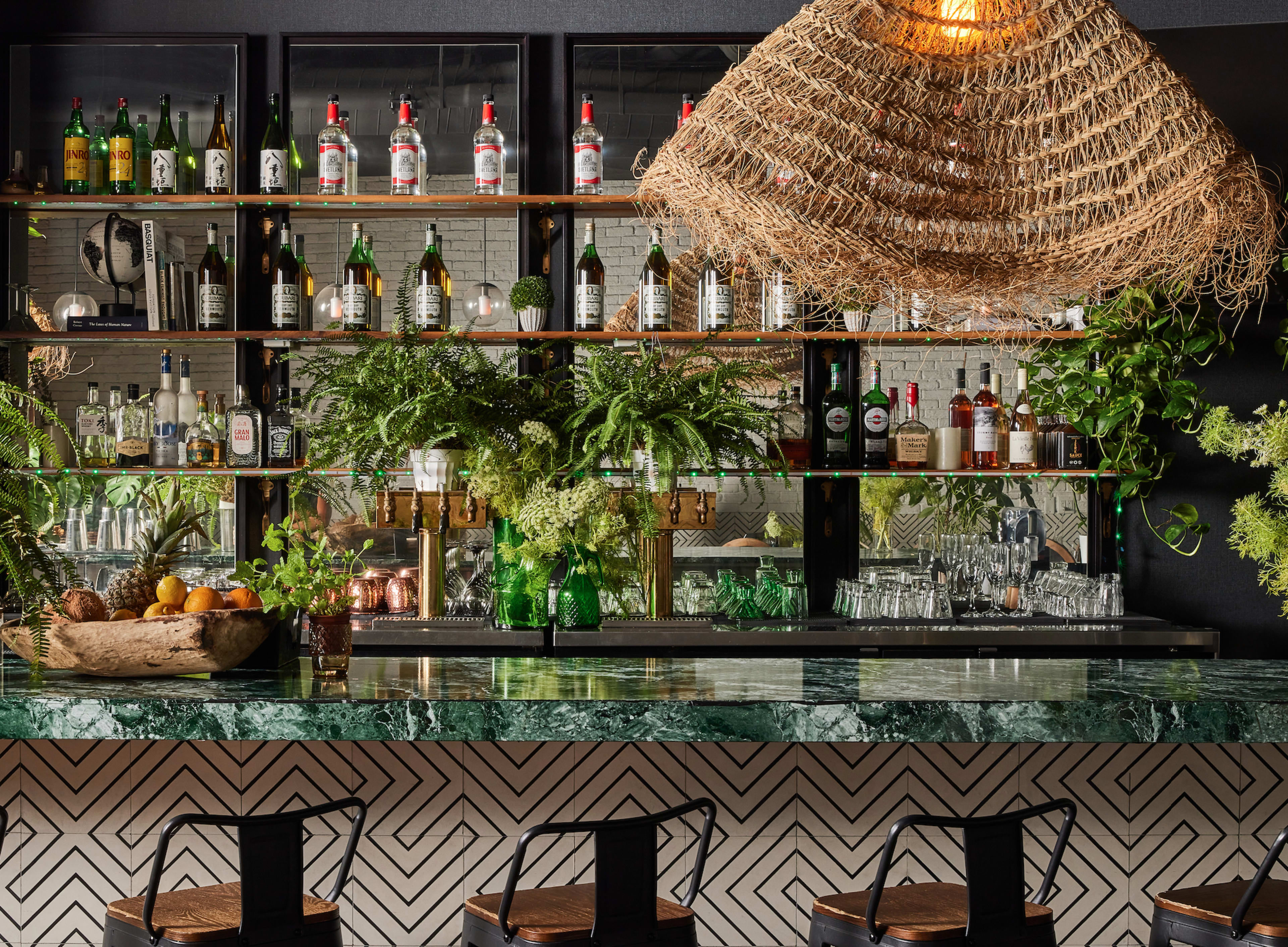 A modern bar features a green marble countertop, a variety of liquor bottles displayed on shelves behind it, and decorative plants throughout the space.