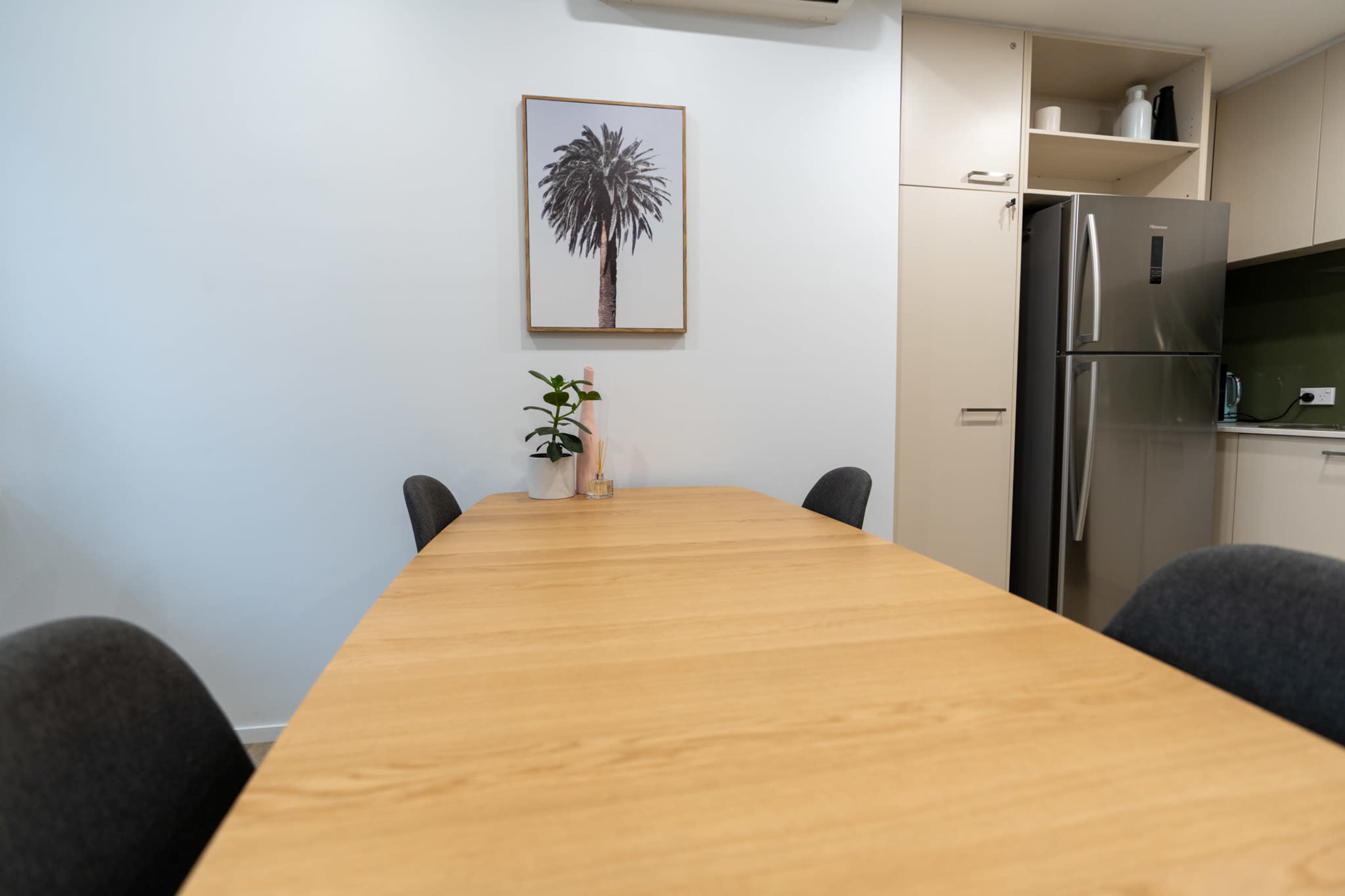 A wooden dining table is set in a well-lit kitchen area, with a palm tree print on the wall and a refrigerator in the background.