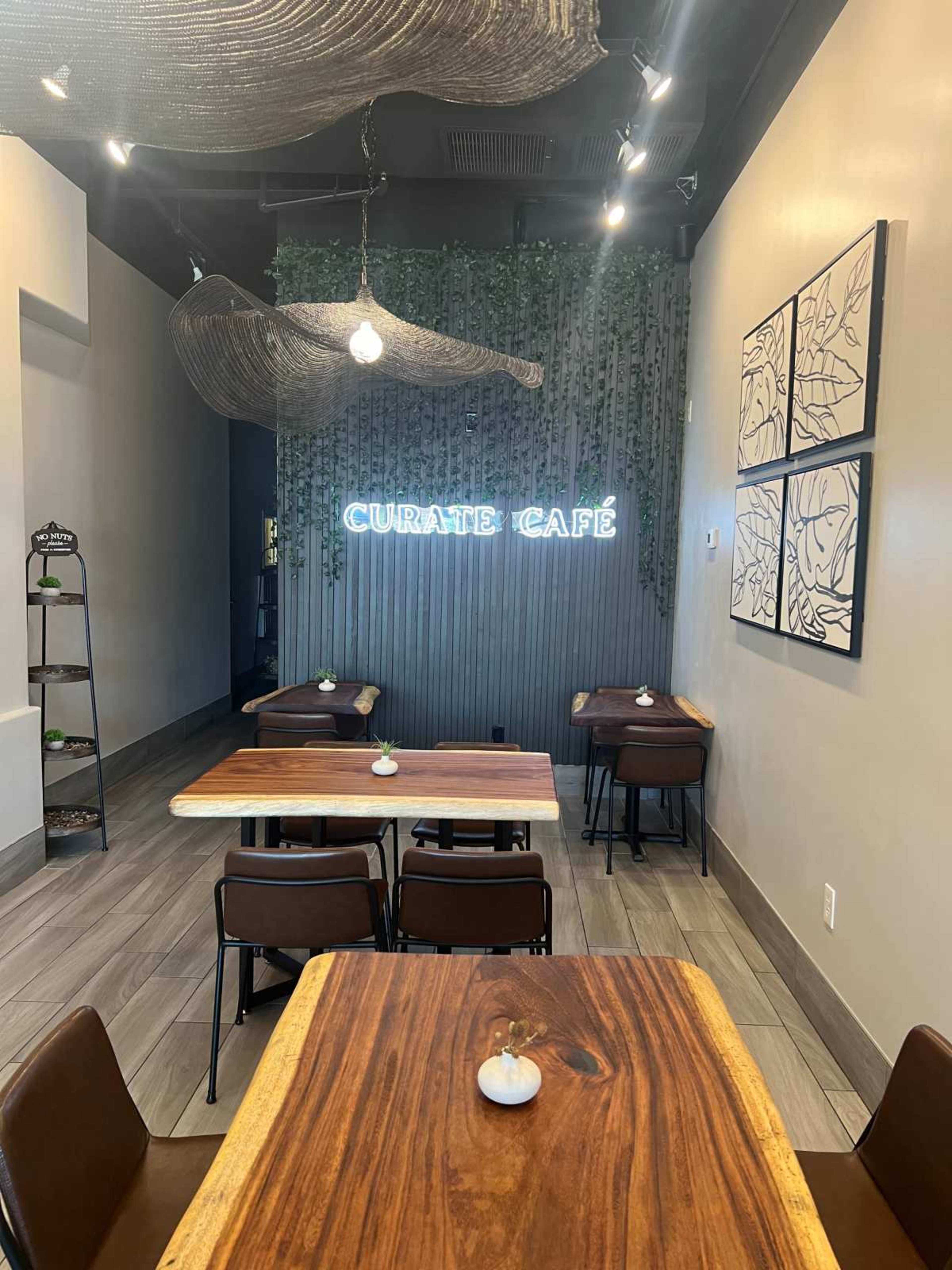 The image shows a small café interior with wooden tables, brown chairs, and a neon sign that reads "CURATE CAFÉ" against a green wall adorned with plants.