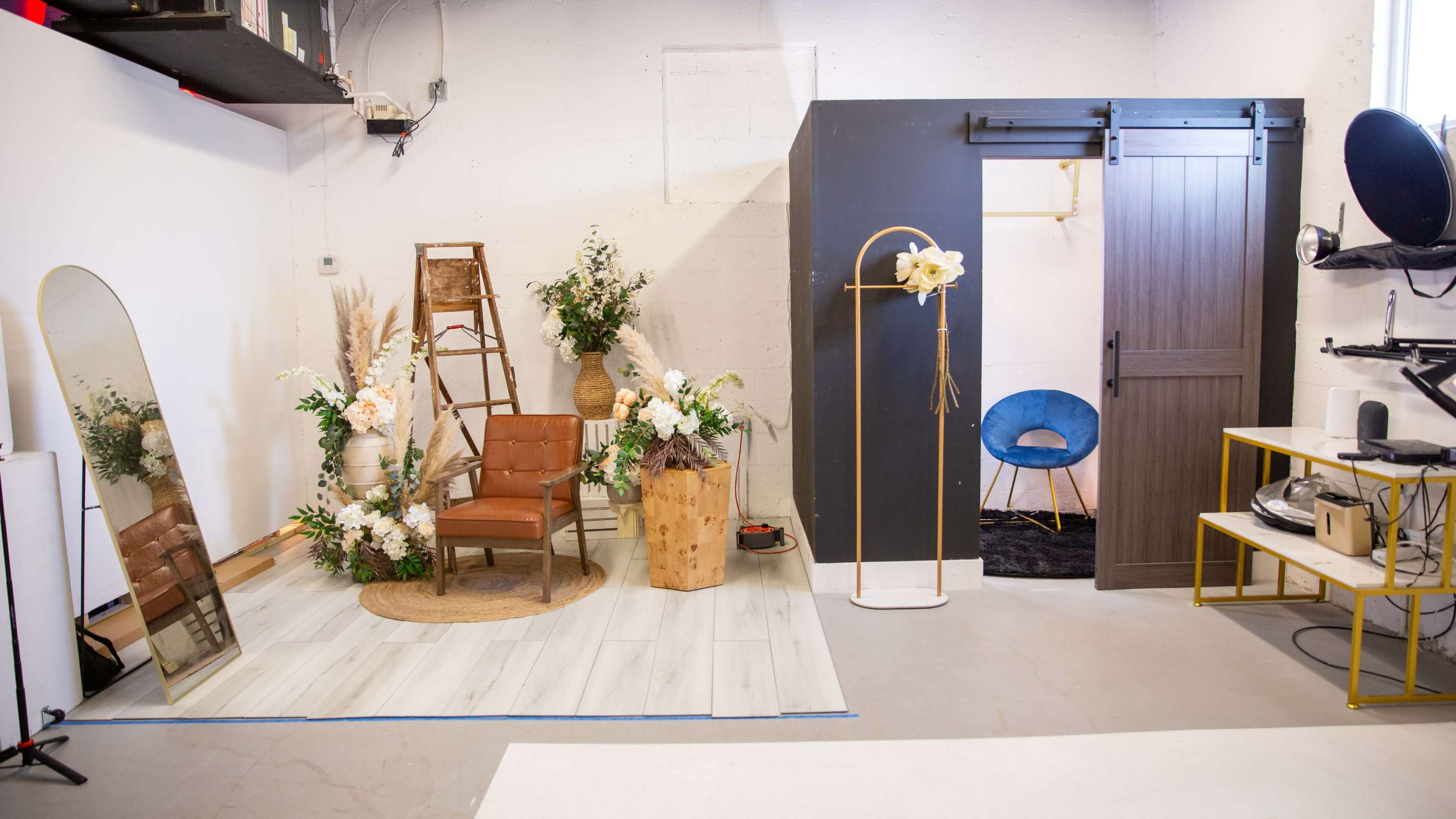 The image shows a small studio space with a decorated area featuring a brown chair, a ladder, and decorative plants, alongside a dark partition with a seating area and various furnishings.