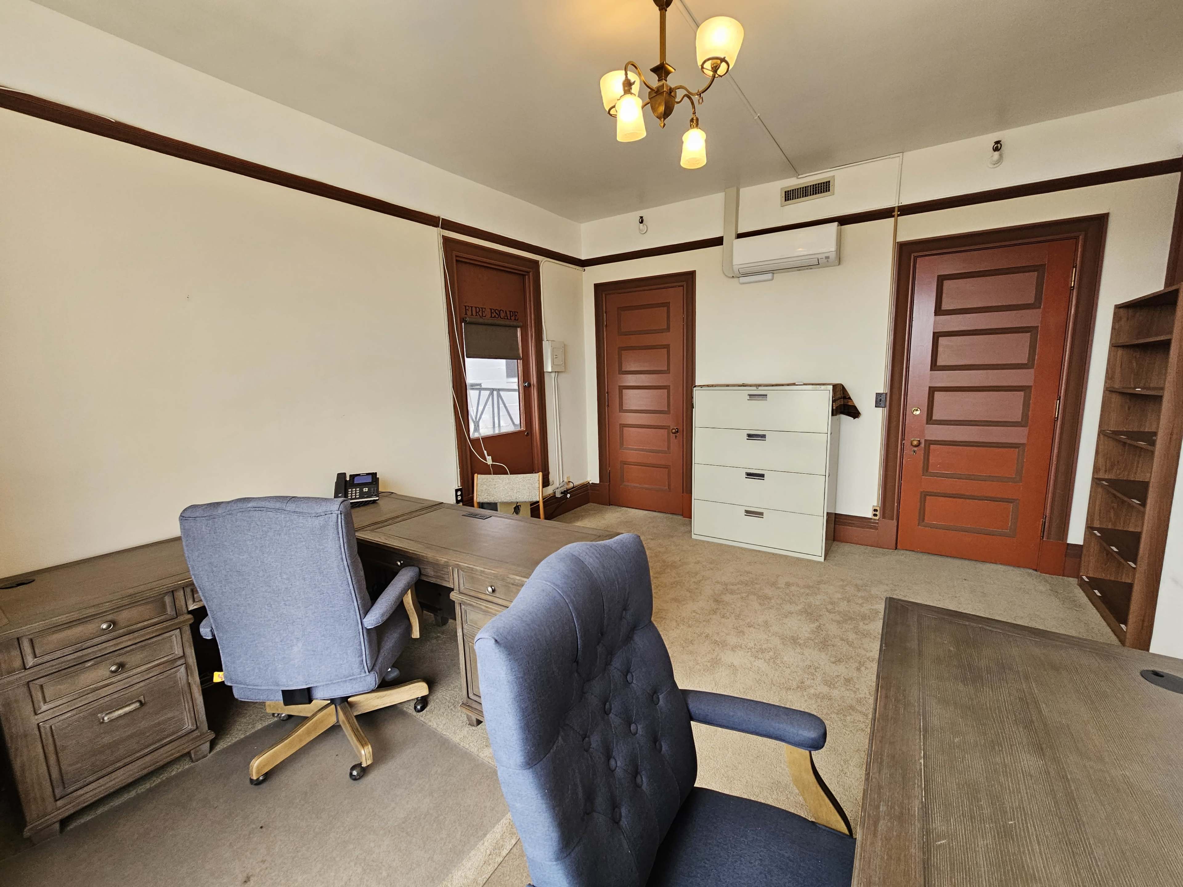 A spacious office with two desks, two chairs, a filing cabinet, and two doors, all within a room featuring light-colored walls and carpet.