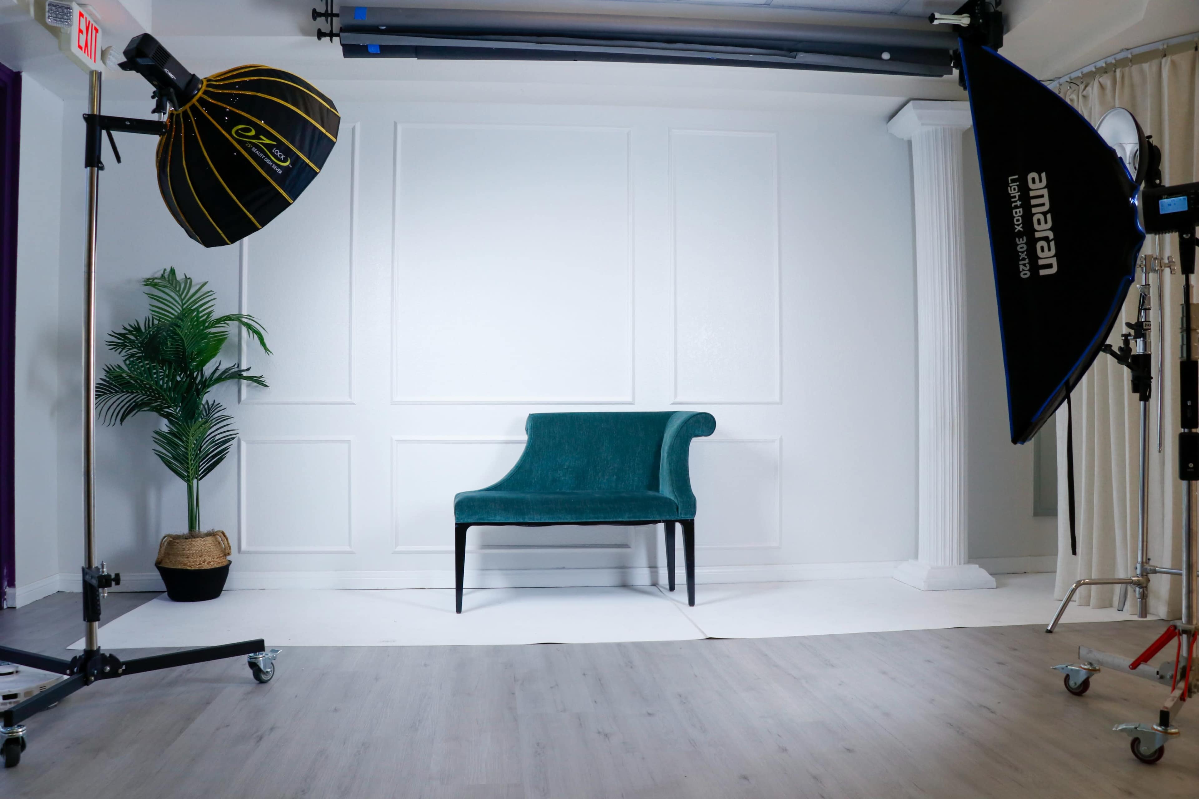 The image shows a photography studio setup with a teal chair positioned in front of a white backdrop, flanked by studio lights and a potted plant.