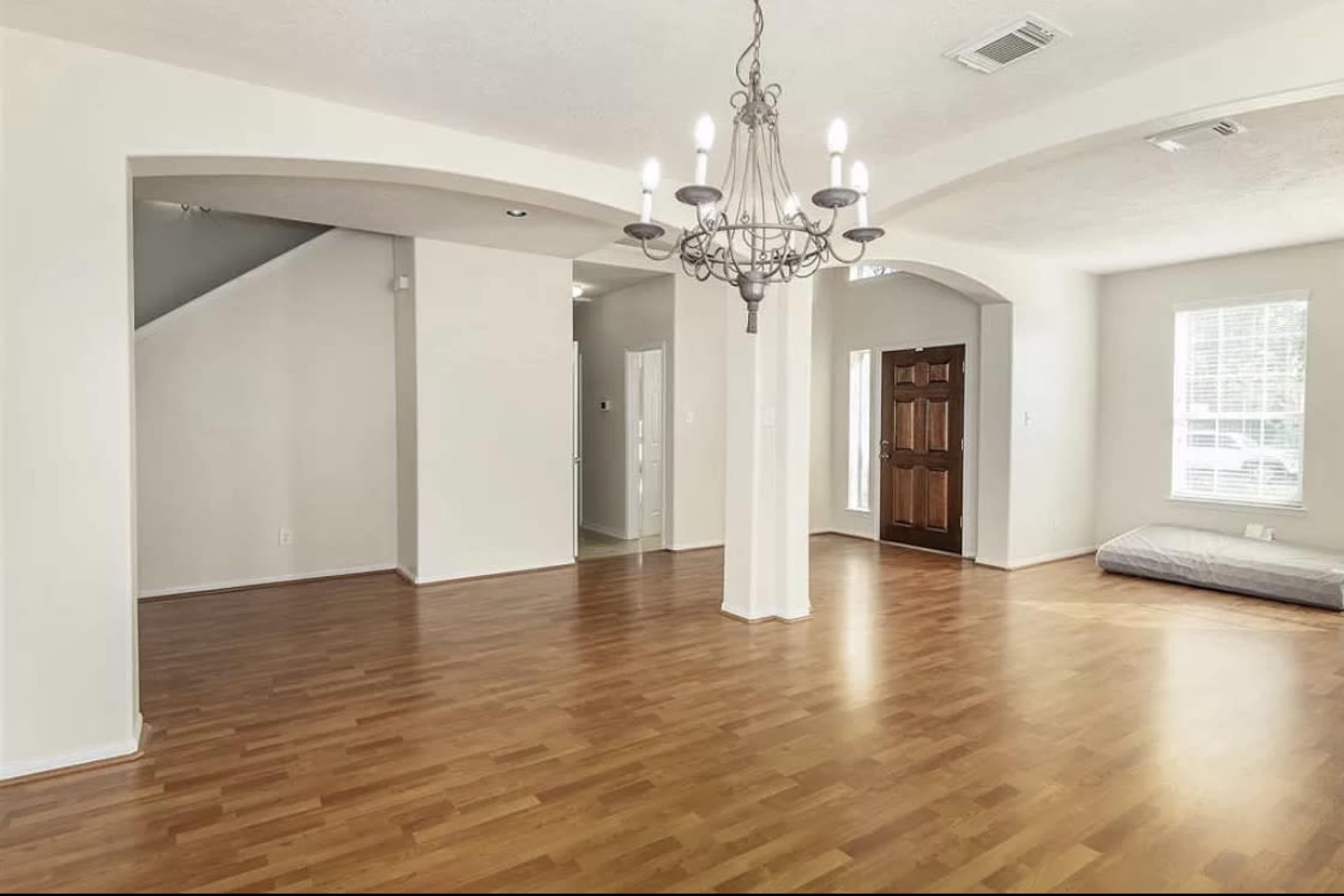 An empty living room with wooden flooring, a chandelier hanging from the ceiling, and an arched entrance leading to another room.