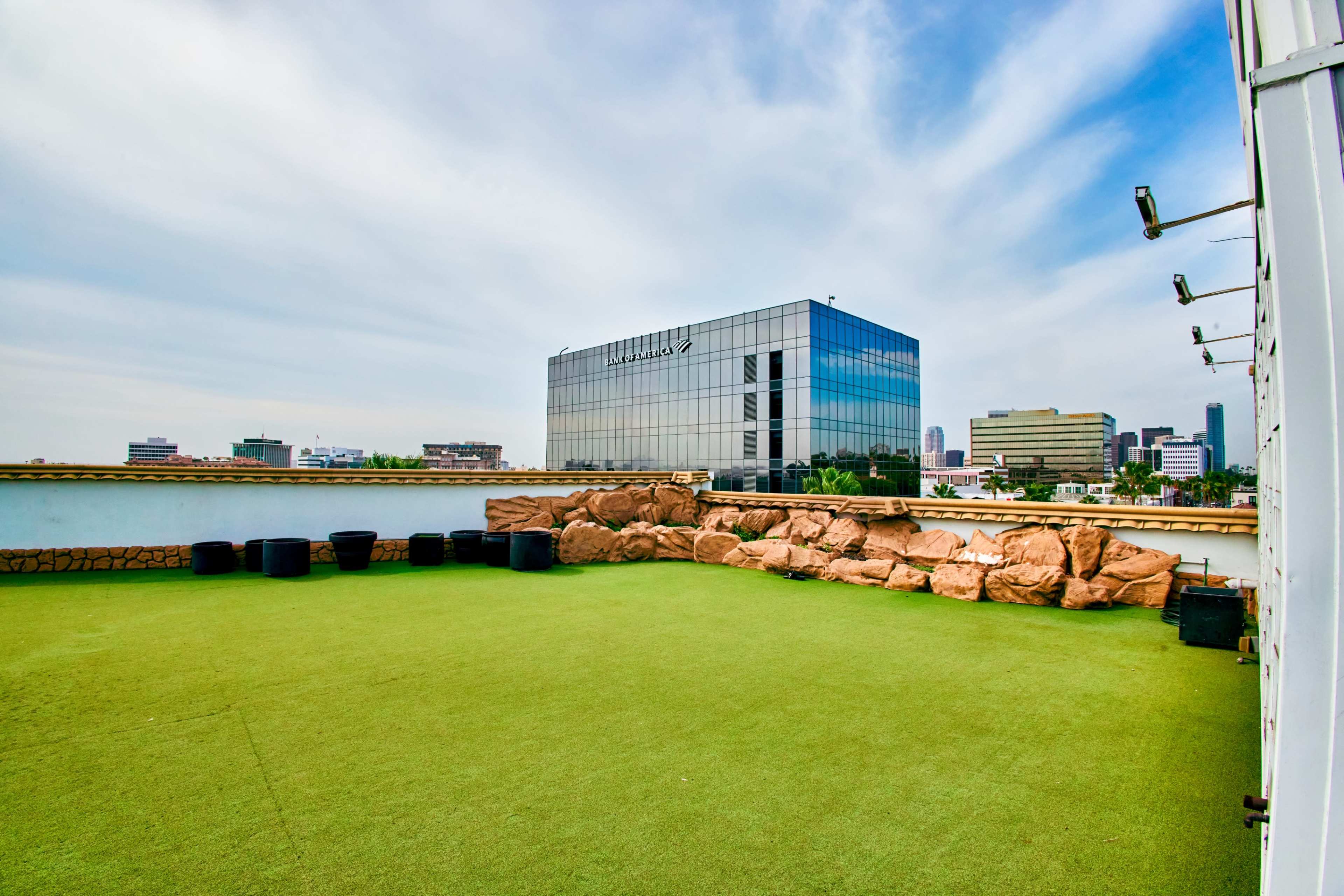 The image shows a green rooftop area with artificial grass and large rocks, overlooking a modern glass building and a city skyline in the background.