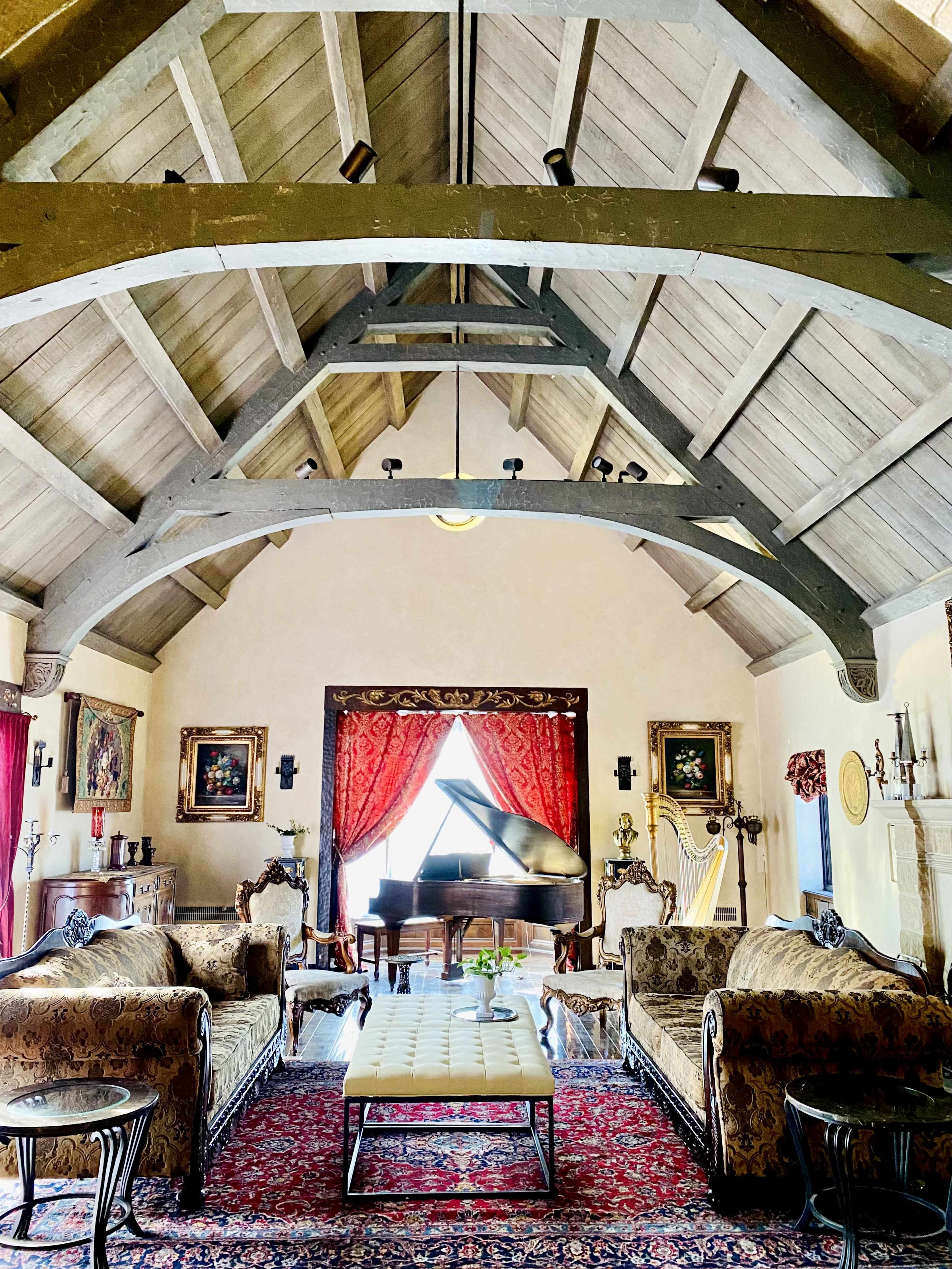 The image shows a spacious, elegantly furnished room featuring a grand piano, ornate furniture, and high wooden beams supporting the ceiling.