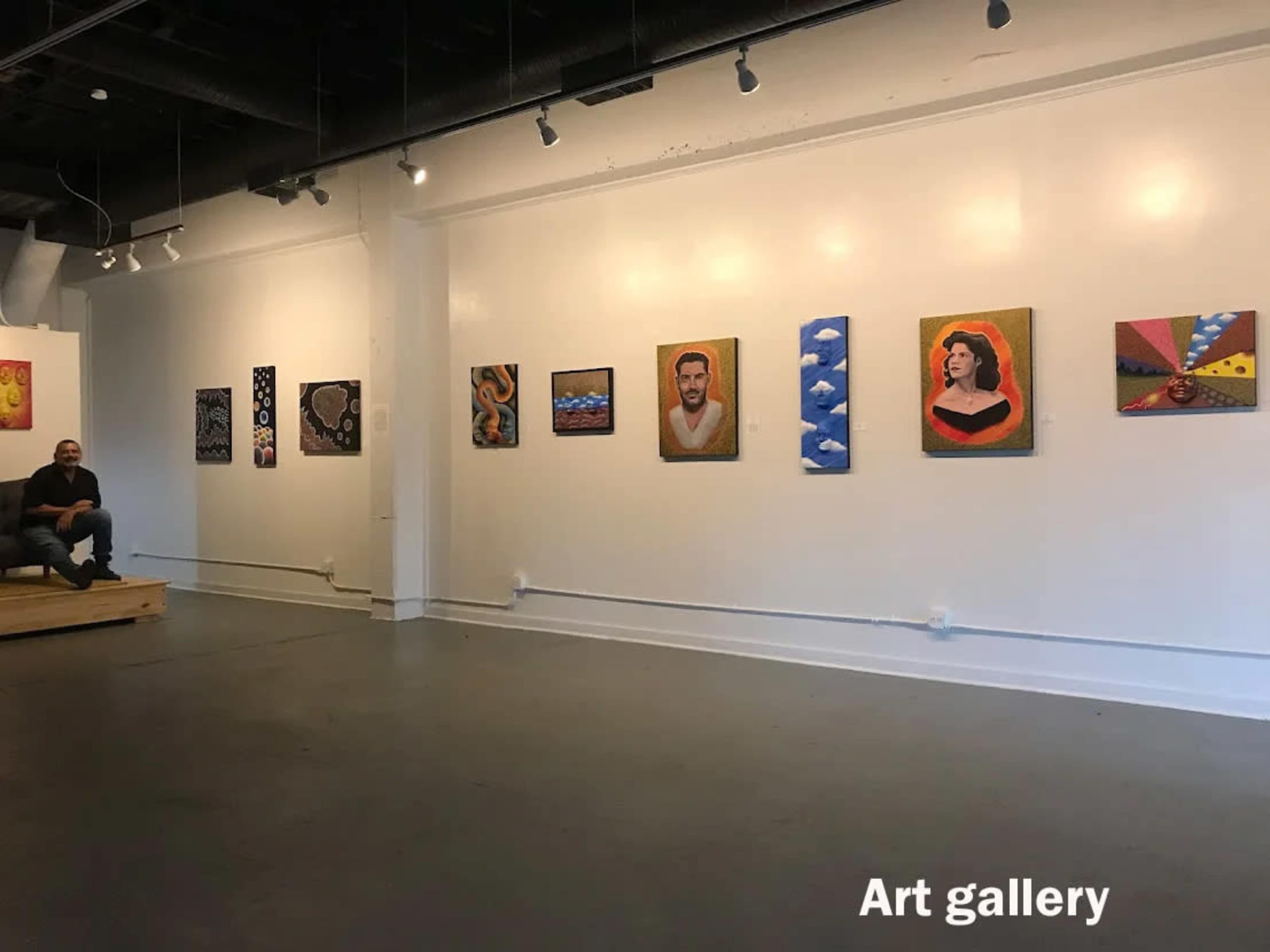 An art gallery features a series of colorful paintings displayed on white walls, with a person seated on a wooden platform.