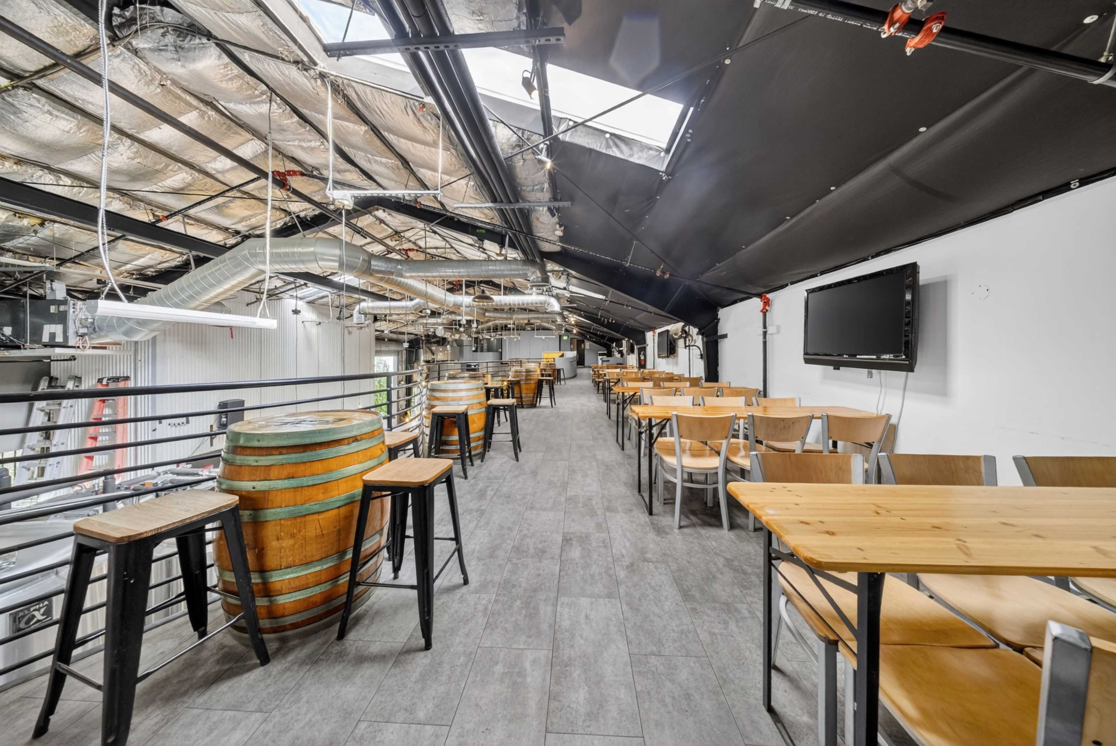 The image shows a spacious, industrial-style dining area with wooden tables, high stools, and decorative barrels along the sides.