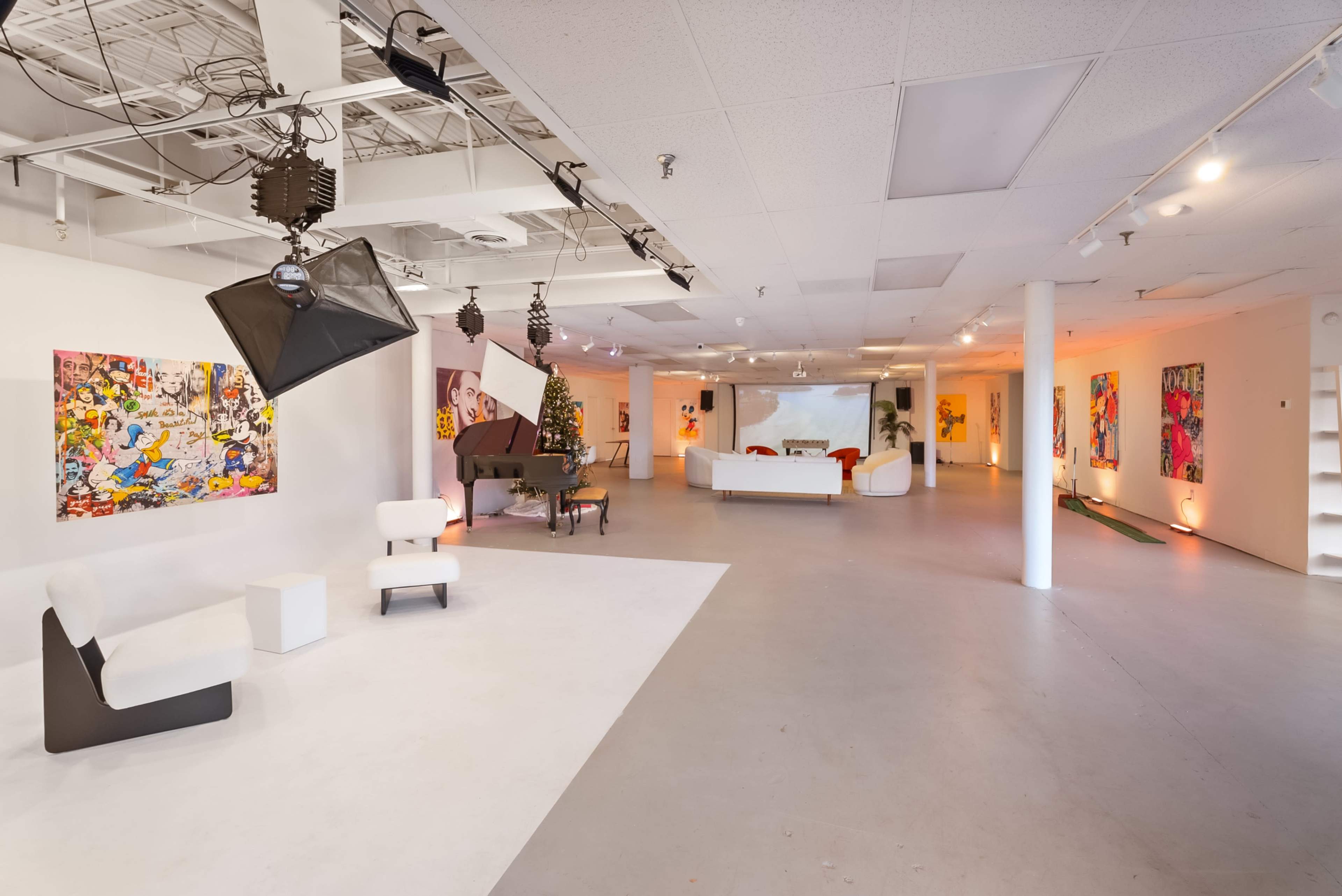 The image shows a spacious, brightly lit art gallery featuring white walls adorned with colorful paintings and contemporary furniture arranged throughout the room.