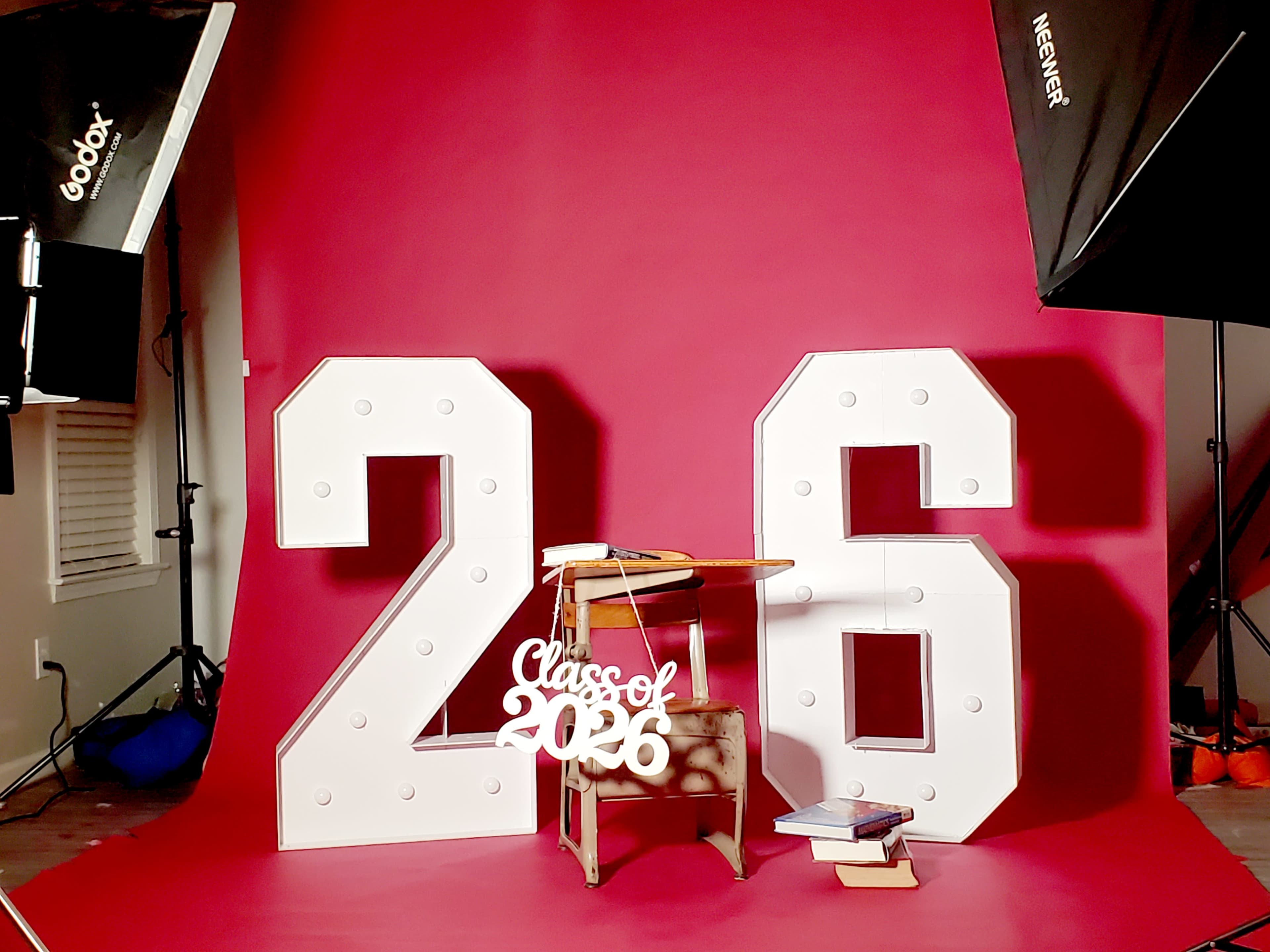 The image features large, white numbers "2" and "6" against a pink backdrop, with a small wooden desk displaying a sign that reads "Class of 2026" and some books.