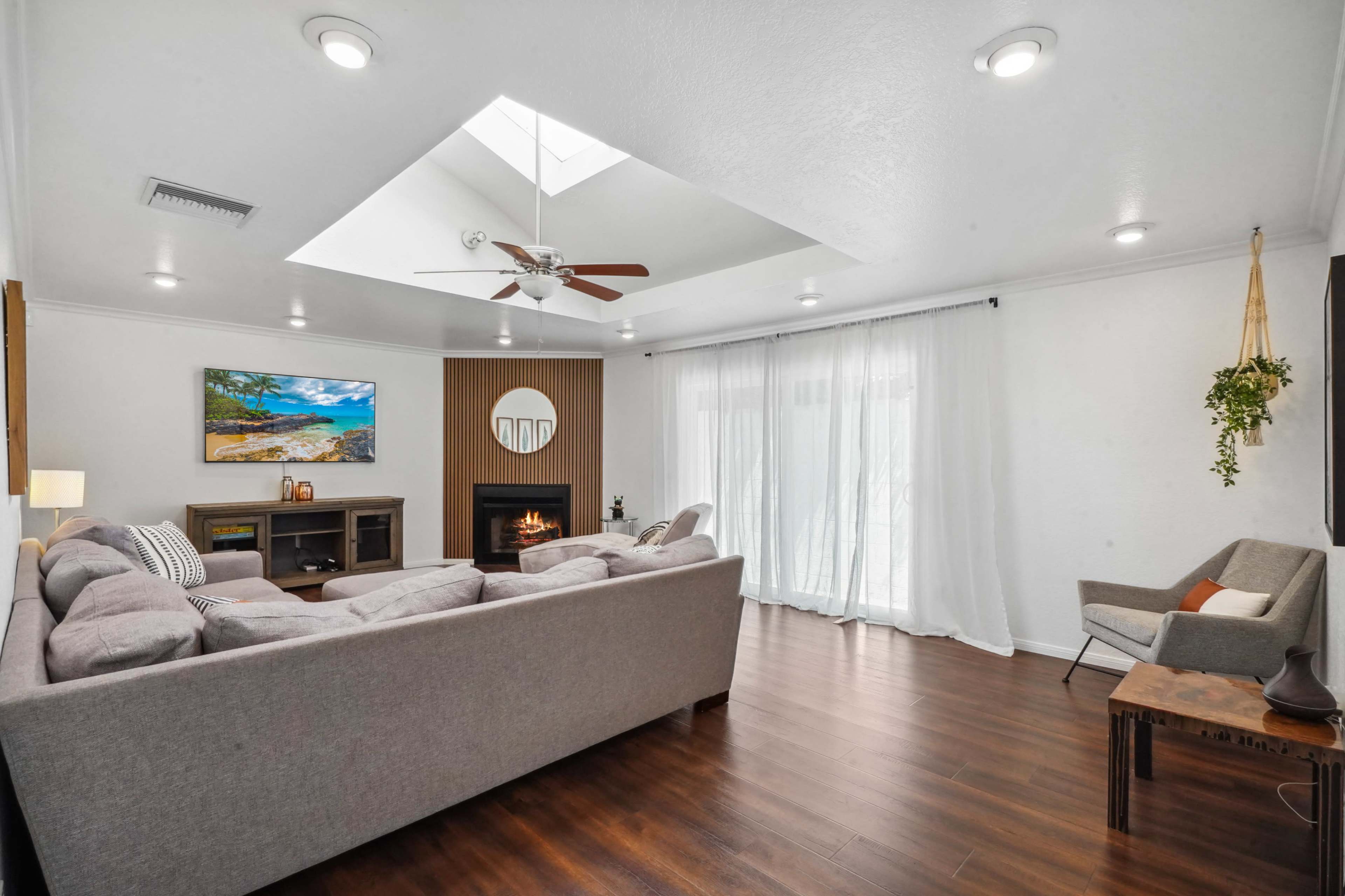 A spacious living room features a light-colored sectional sofa, large windows with sheer curtains, and a modern fireplace.