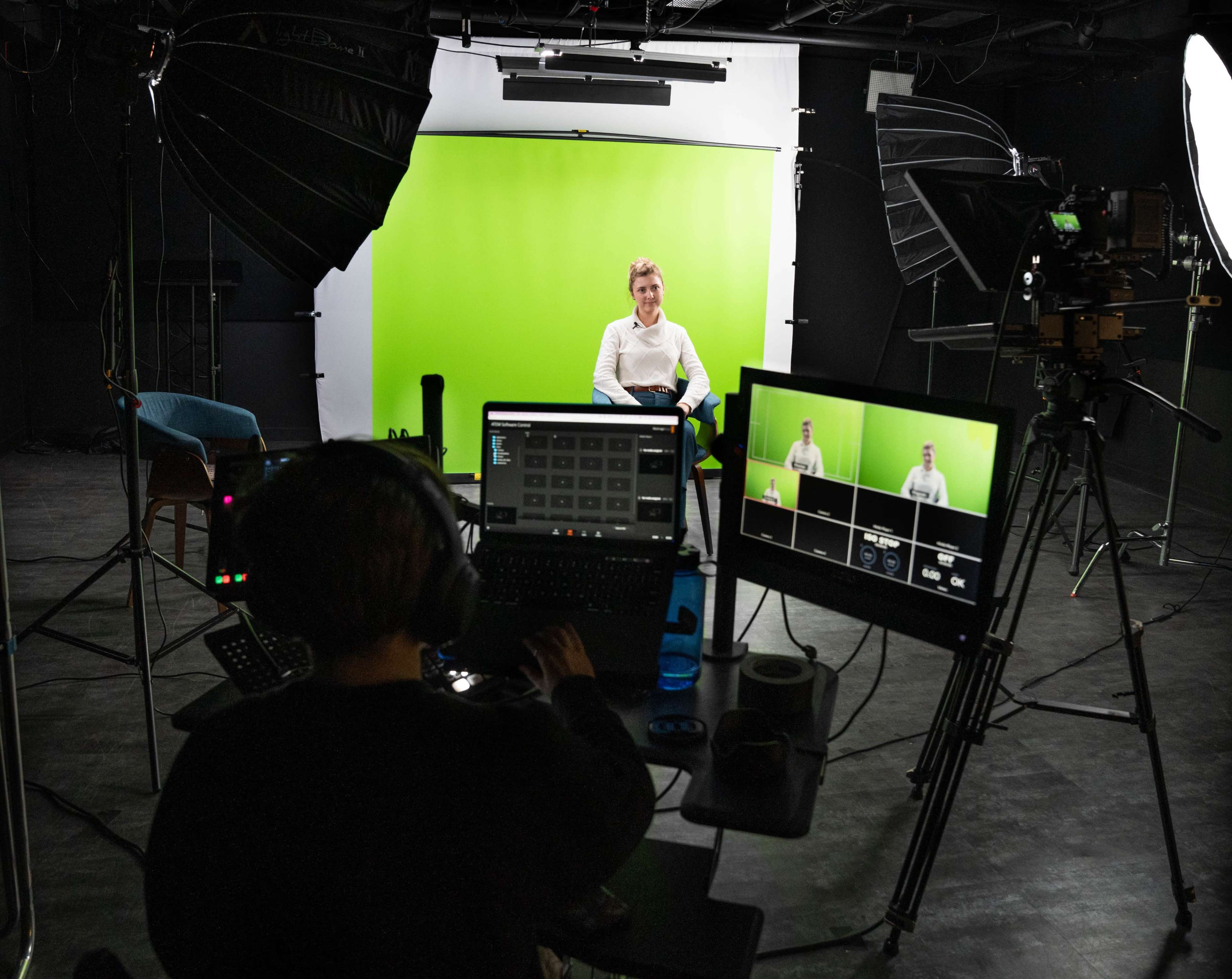 A person is seated in front of a green screen while another individual operates a control panel in a studio setting.