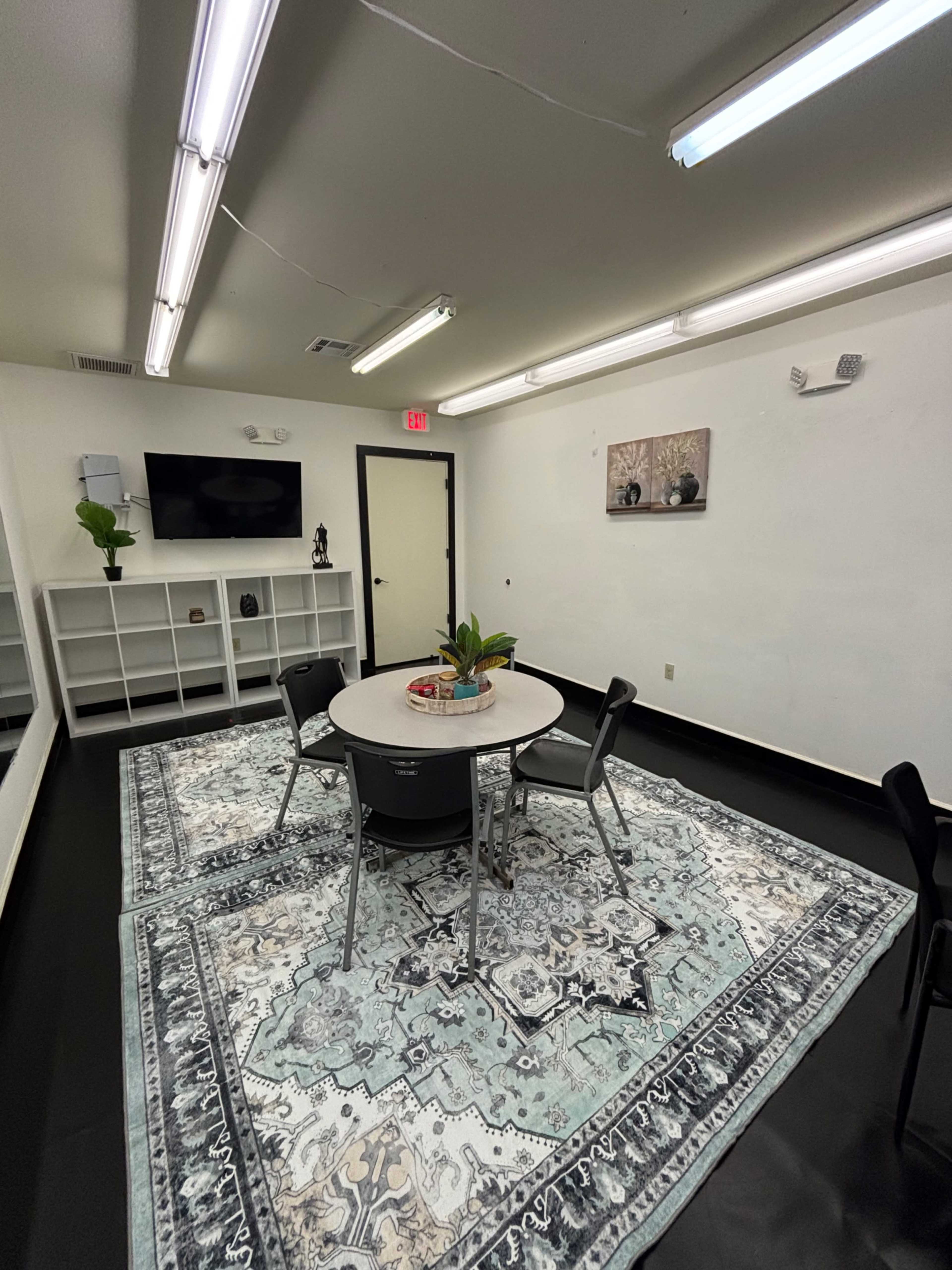 The image shows a small, well-lit room featuring a round table surrounded by four chairs, with a patterned rug on the floor and a wall-mounted TV.