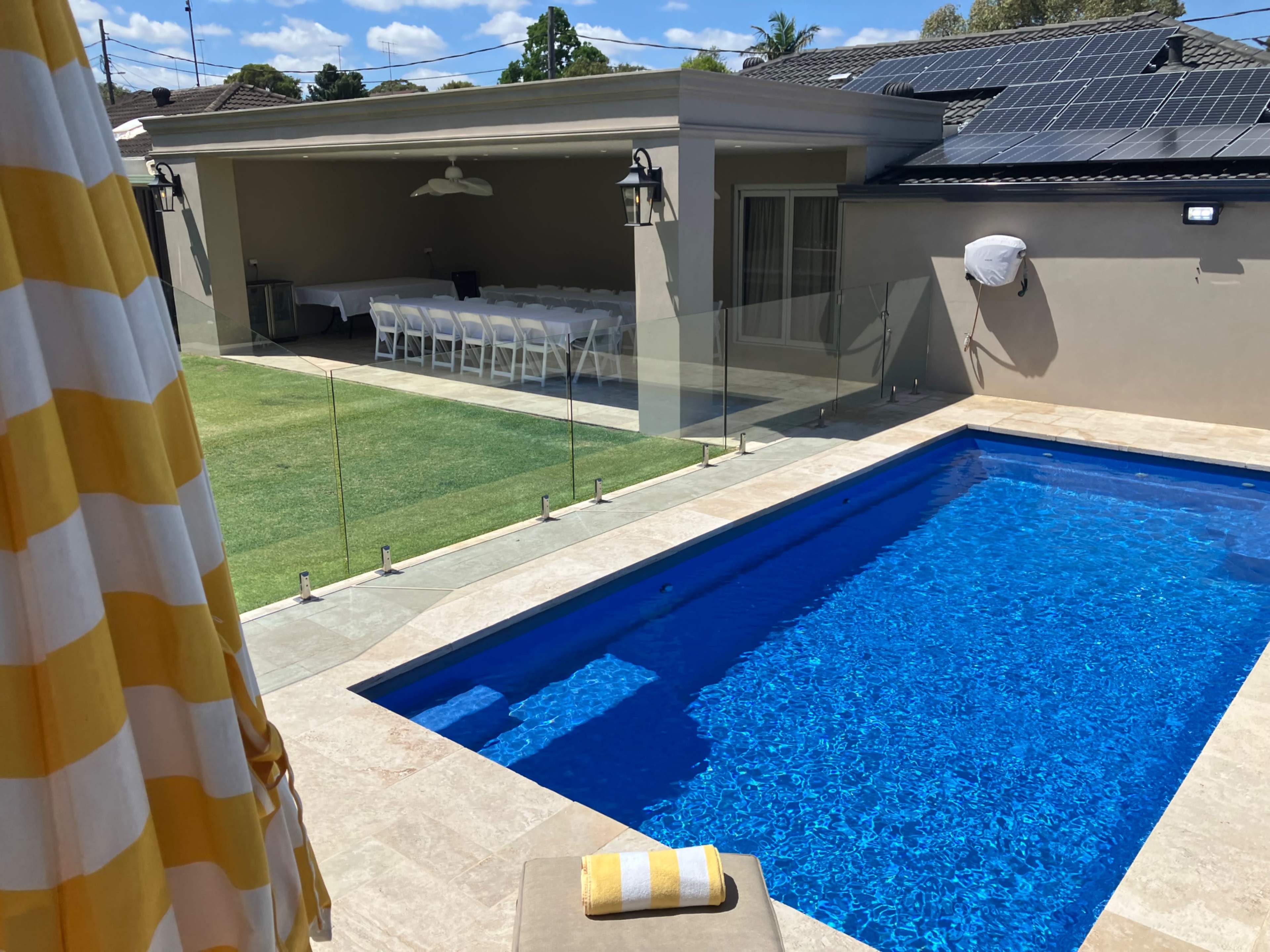 The image shows a backyard with a clear blue swimming pool, a glass fence, and a dining area under a shaded patio.