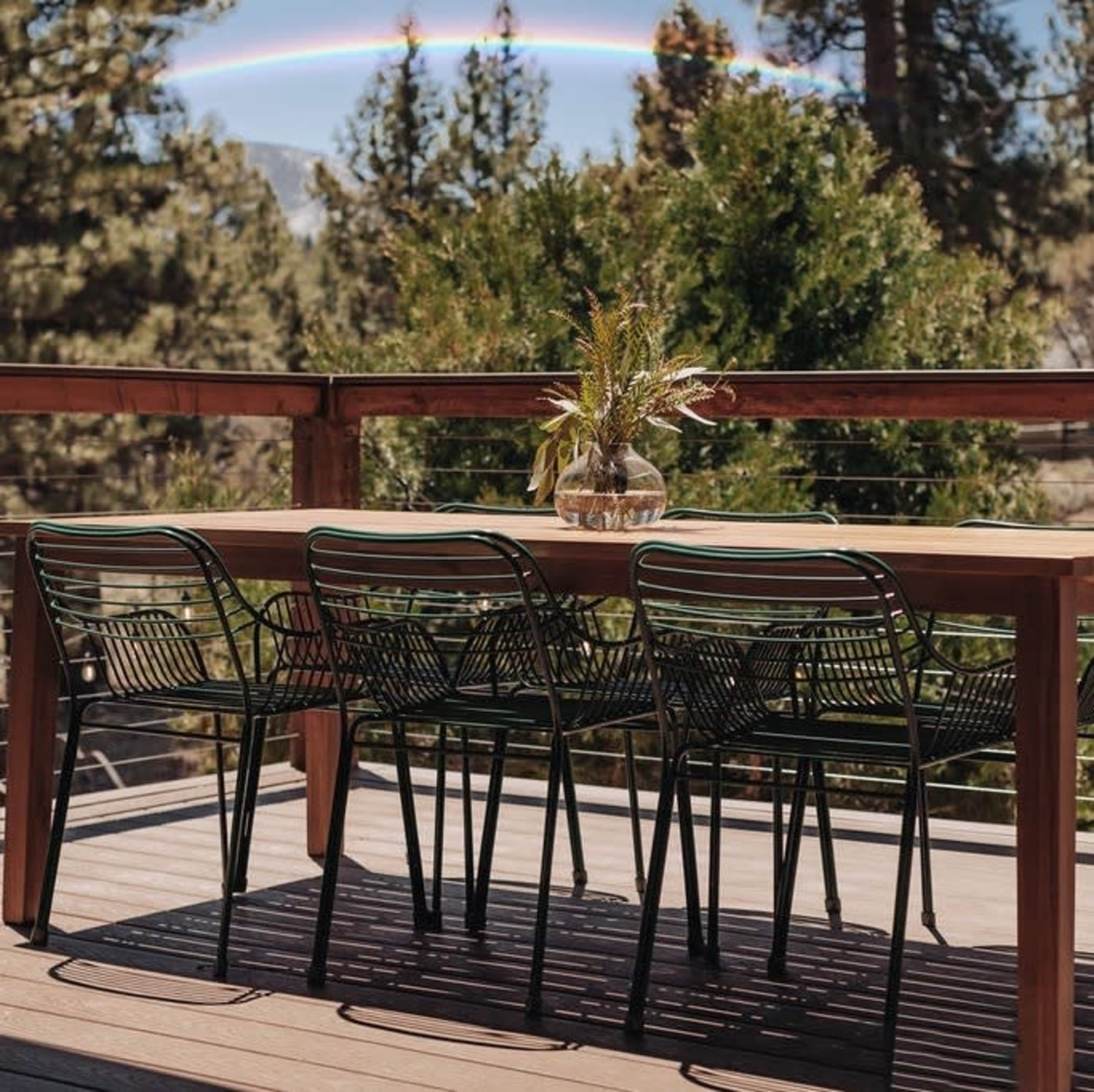 A wooden dining table with black wire chairs is set on a deck overlooking trees and a rainbow in the sky.