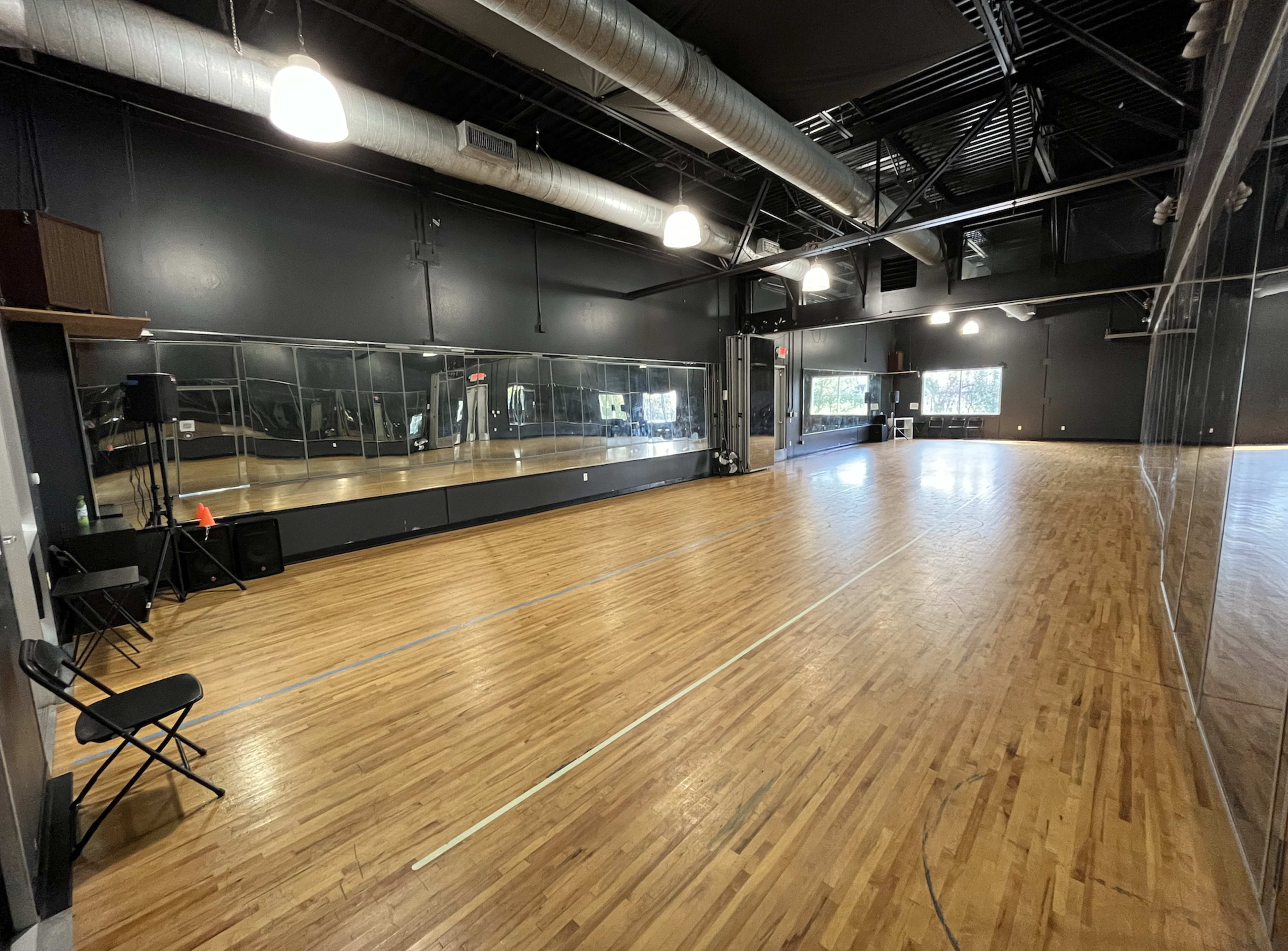 A spacious dance studio with wooden floors, black walls, and mirrors lining one side.