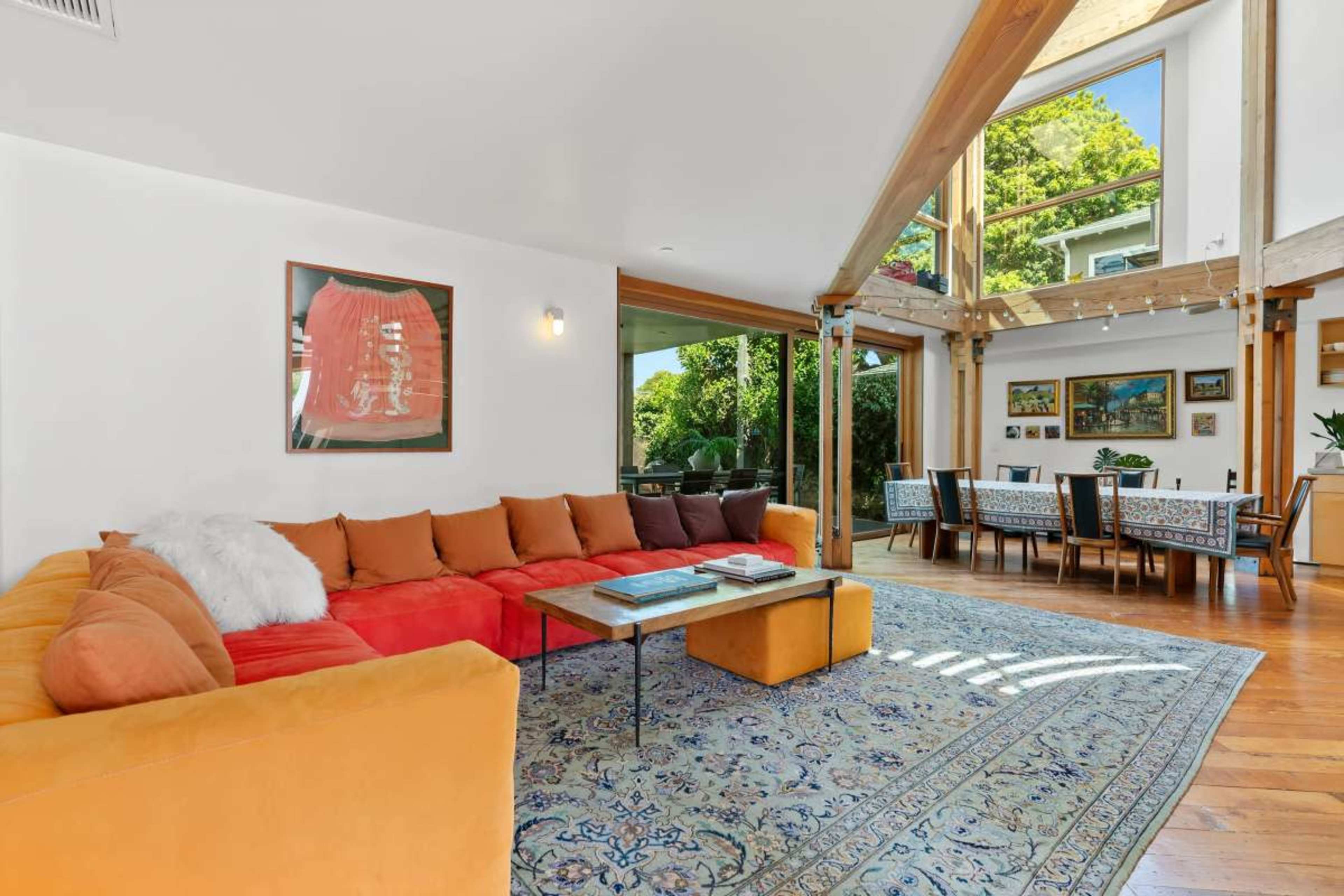 The image shows a modern living room with a large orange sectional sofa, a coffee table, a patterned rug, and a dining area visible through large windows.