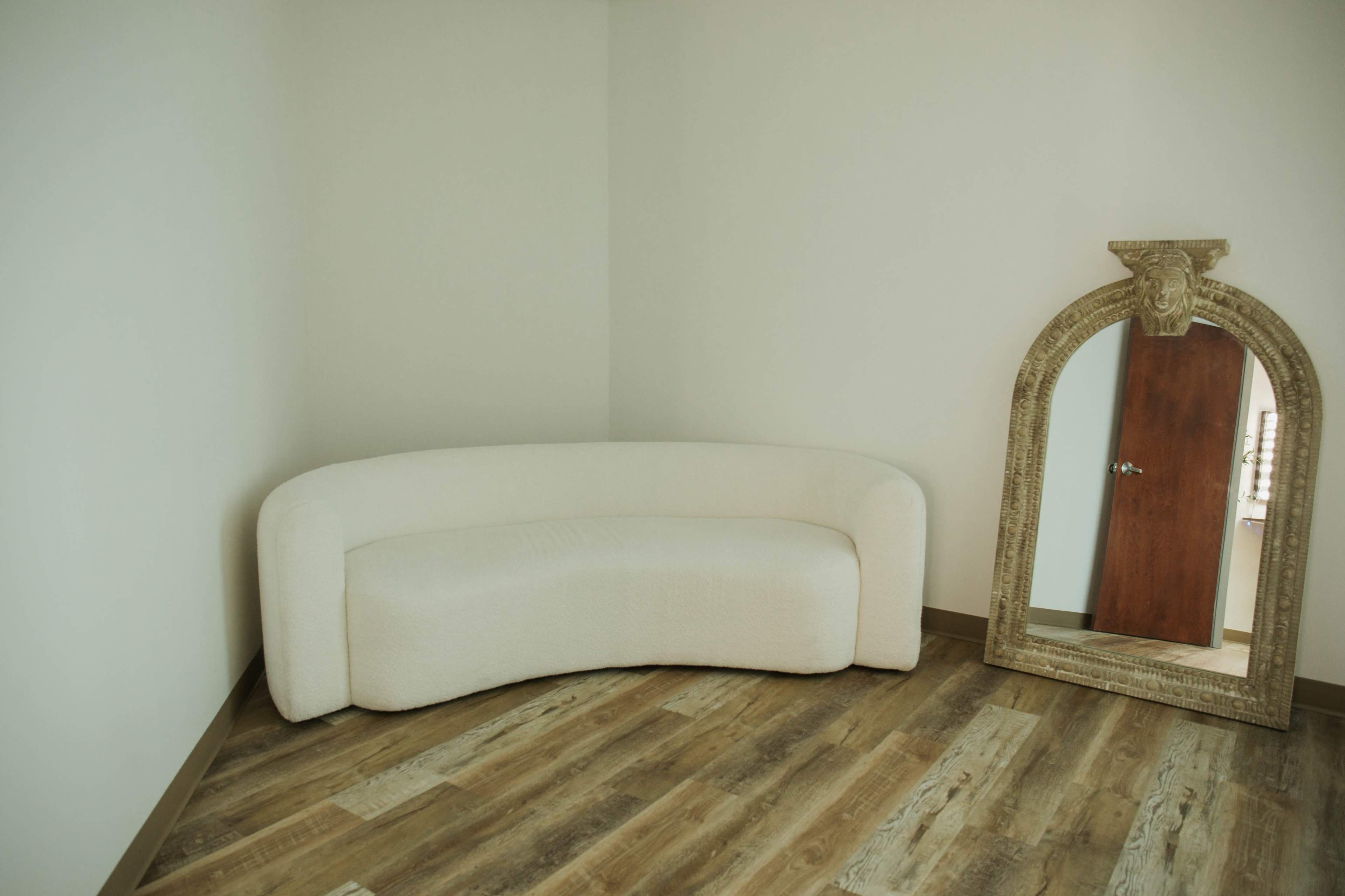 A curved white sofa positioned in a corner of a room with wooden flooring, next to an ornate mirror.