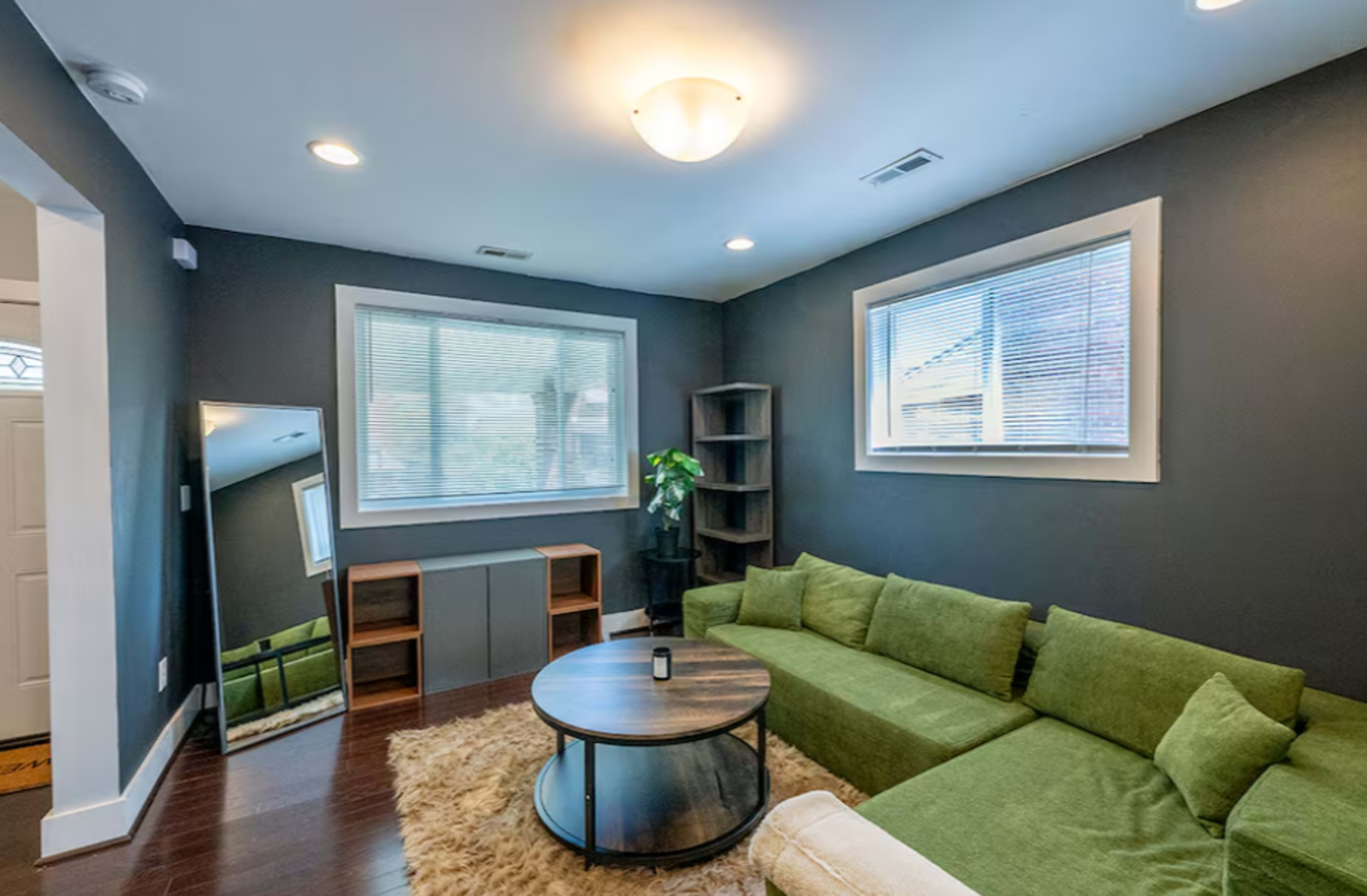 The image shows a modern living room with a green sectional couch, a round coffee table, a mirror, and a bookshelf, all set against dark gray walls.
