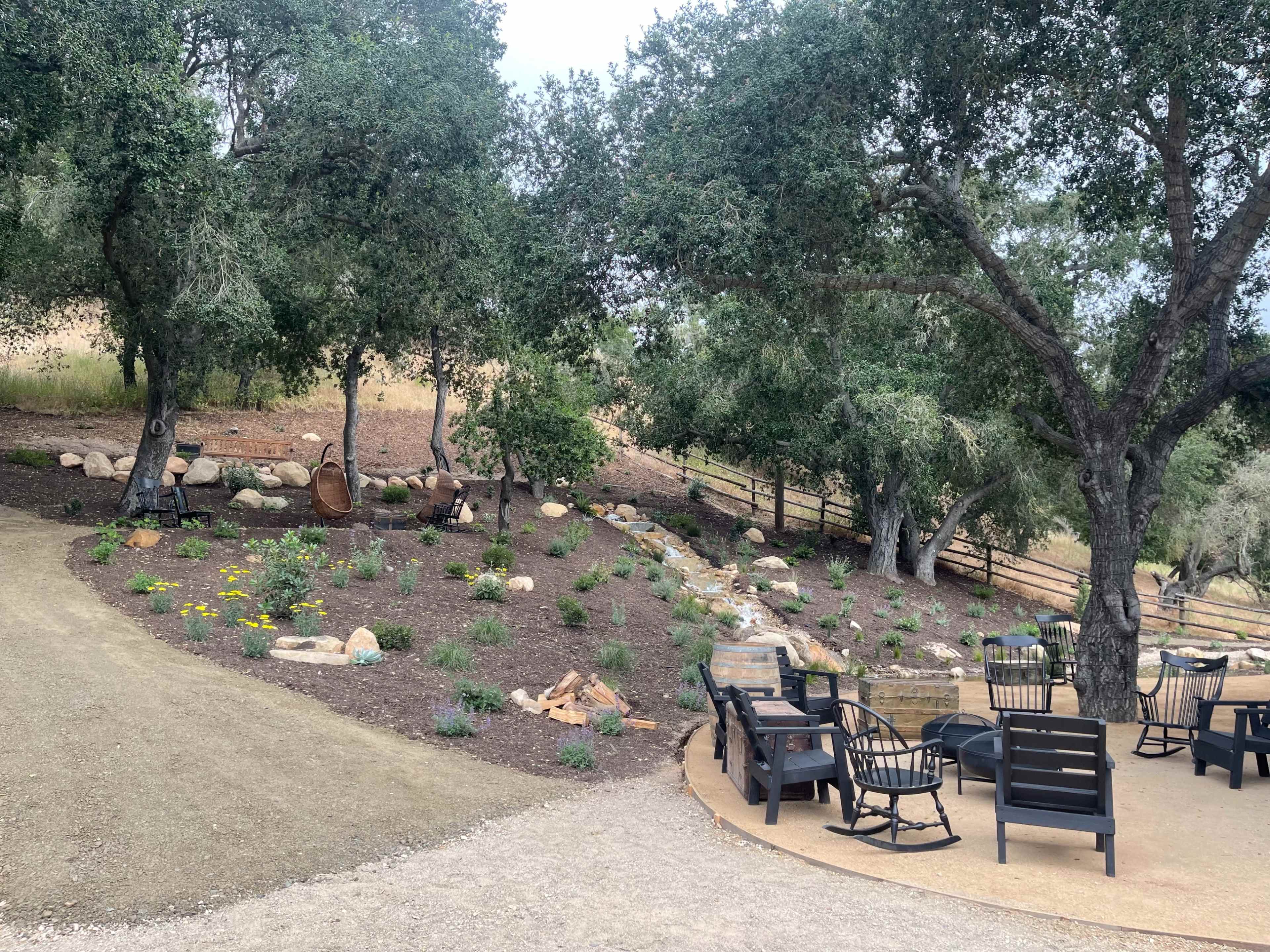 A landscaped area with a gravel path, seating arrangements, and trees, alongside a small stone water feature.