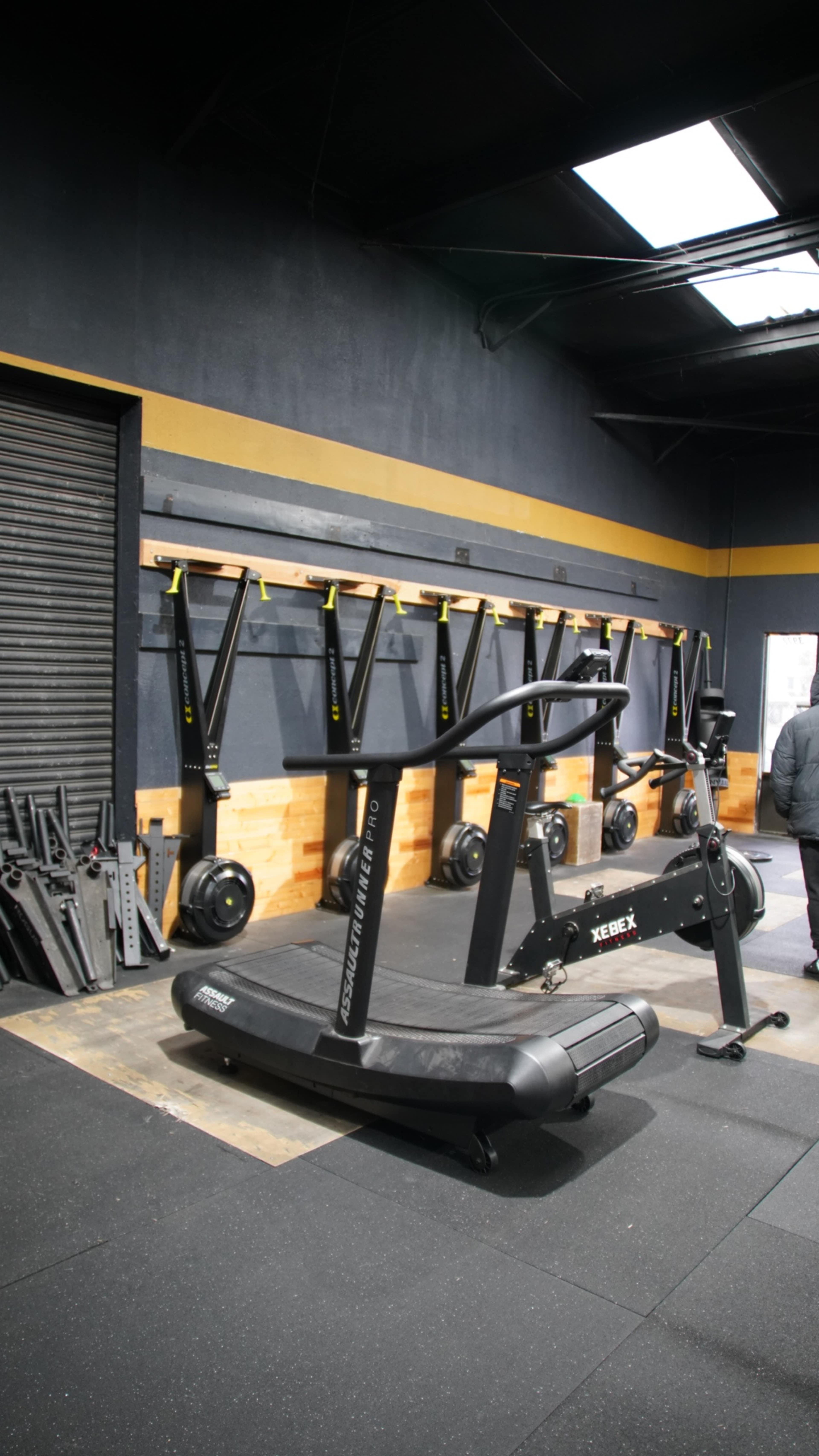 A gym interior featuring a curved treadmill in the foreground and a wall-mounted rack with weights in the background.