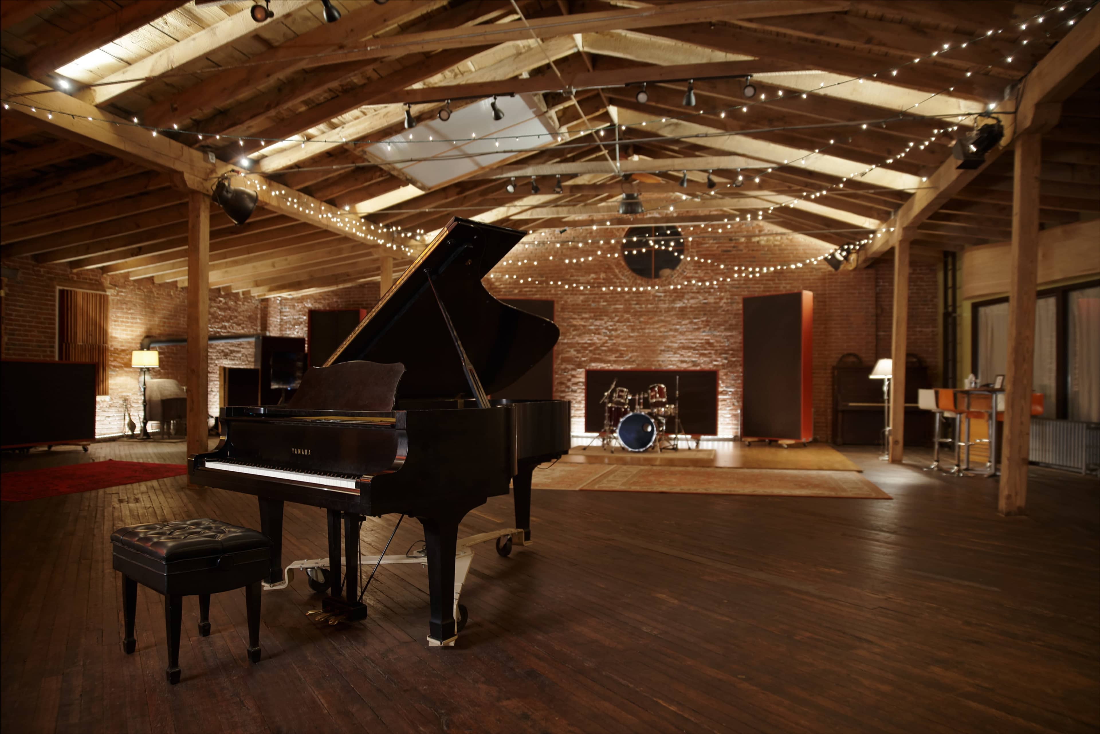 A grand piano is positioned on a wooden floor in a spacious, lit recording studio featuring exposed beams and a small drum set in the background.