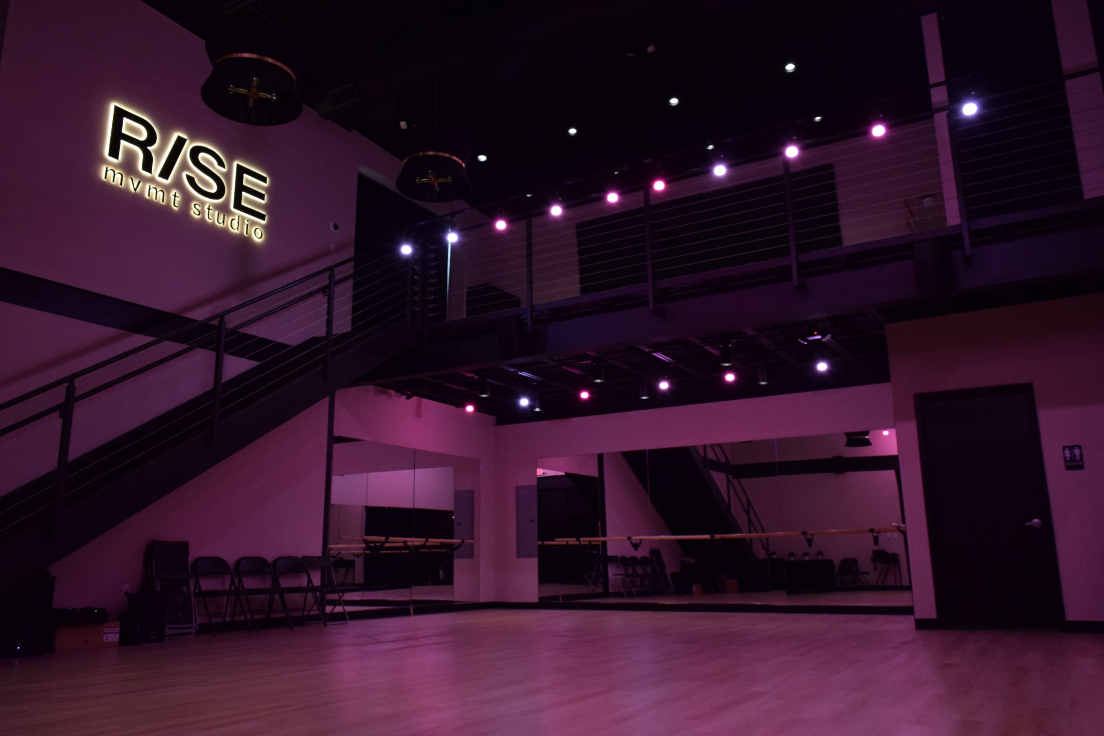 A dance studio with pink and purple lighting, featuring a mirrored wall and a staircase in the background.