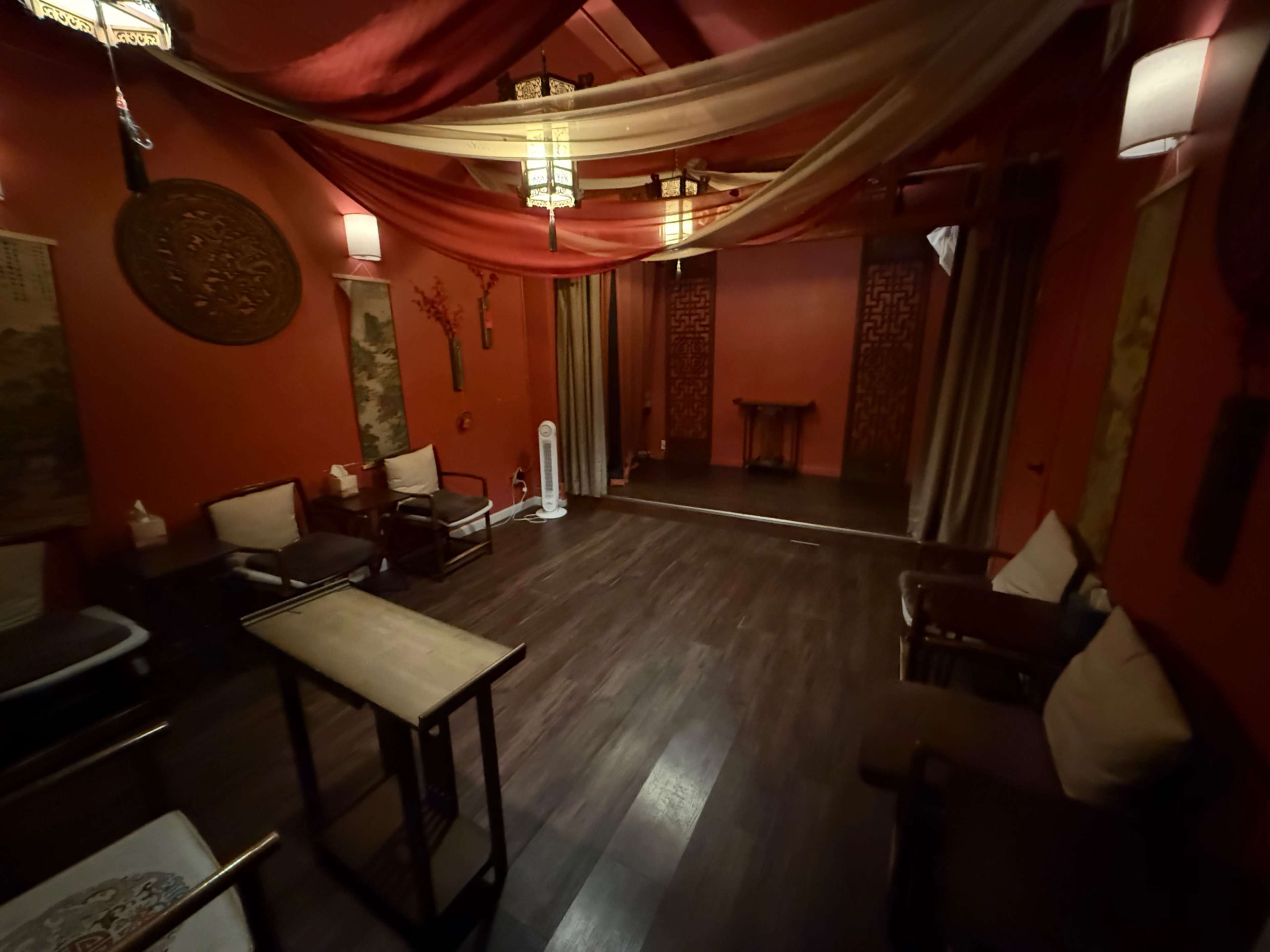The image shows a dimly lit spa or relaxation room with red walls, wooden flooring, and a few chairs arranged around a small table.