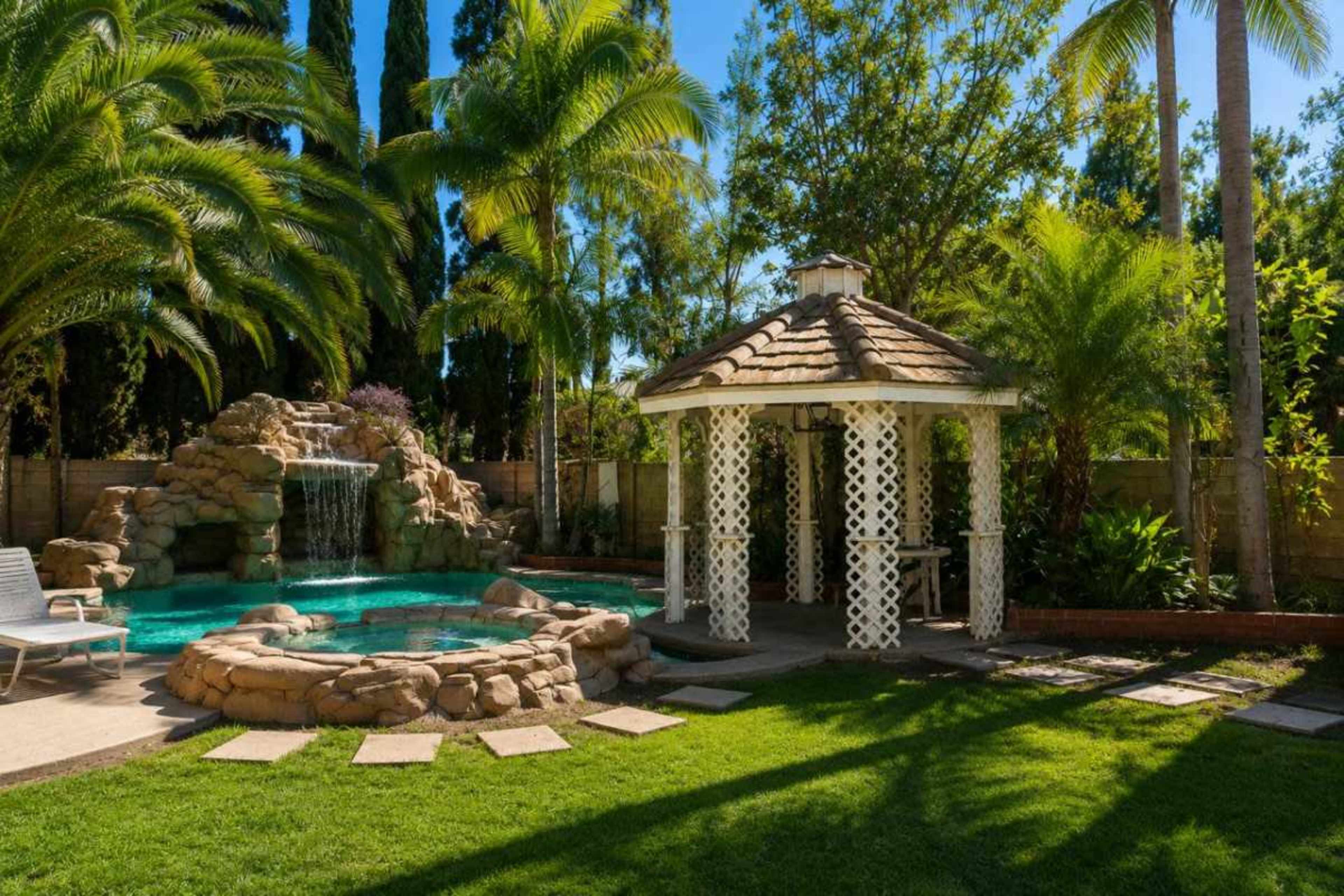 The image shows a landscaped backyard featuring a swimming pool with a rock waterfall, a gazebo, and palm trees surrounding a green lawn.