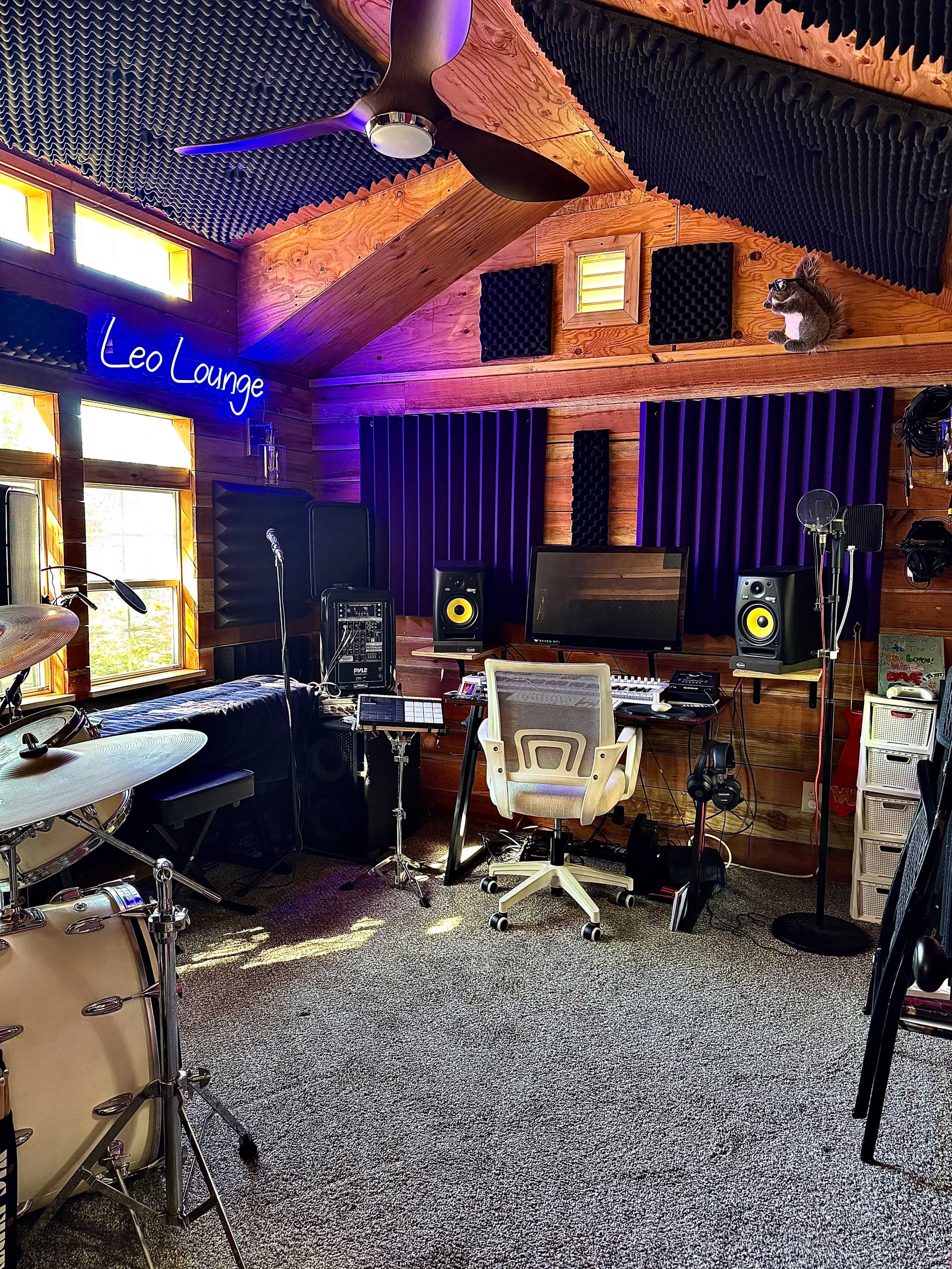 The image shows a music studio with a drum set, keyboard, computer setup, and various audio equipment, all set against a rustic wooden interior.