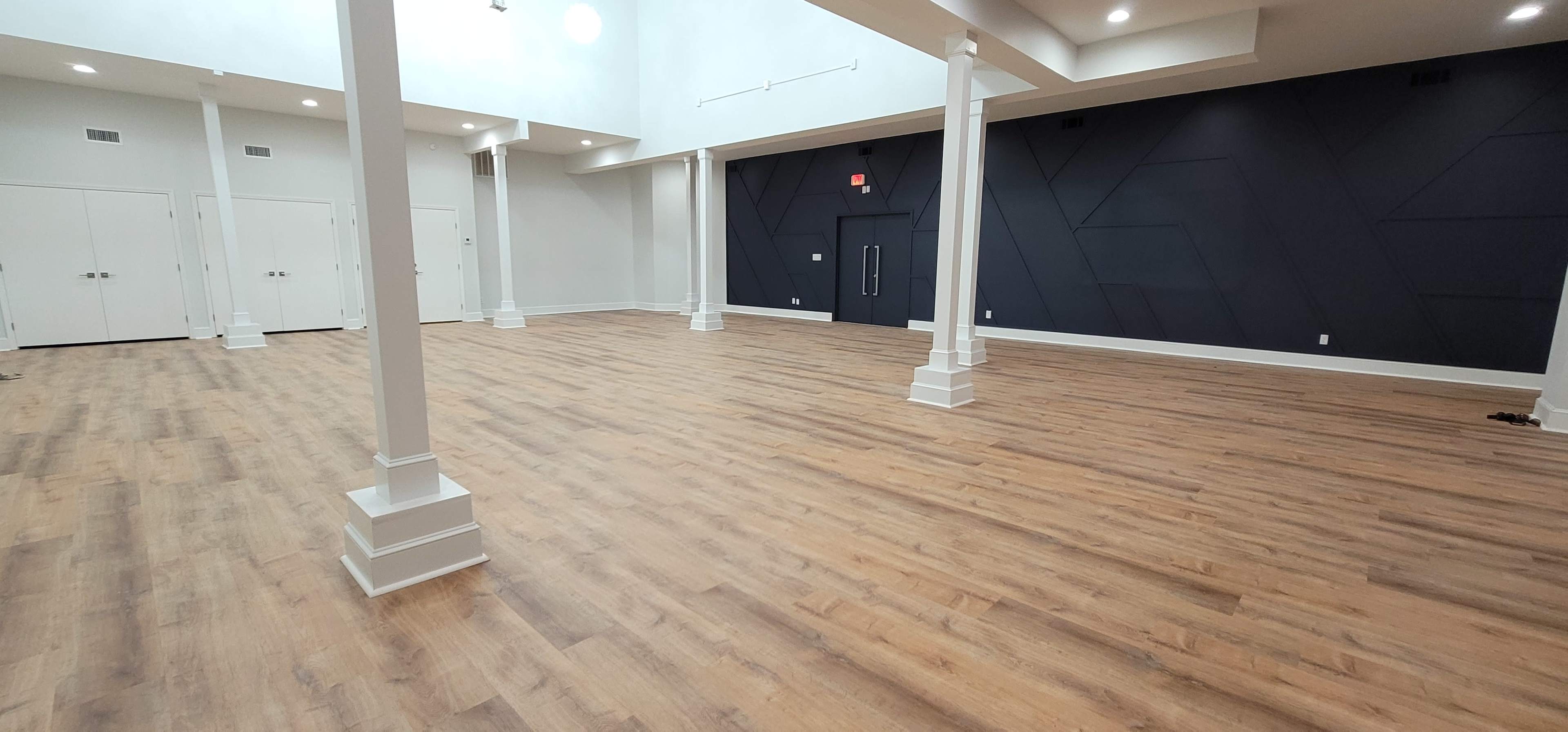 A spacious, empty room with wooden flooring and white columns, illuminated by overhead lights.