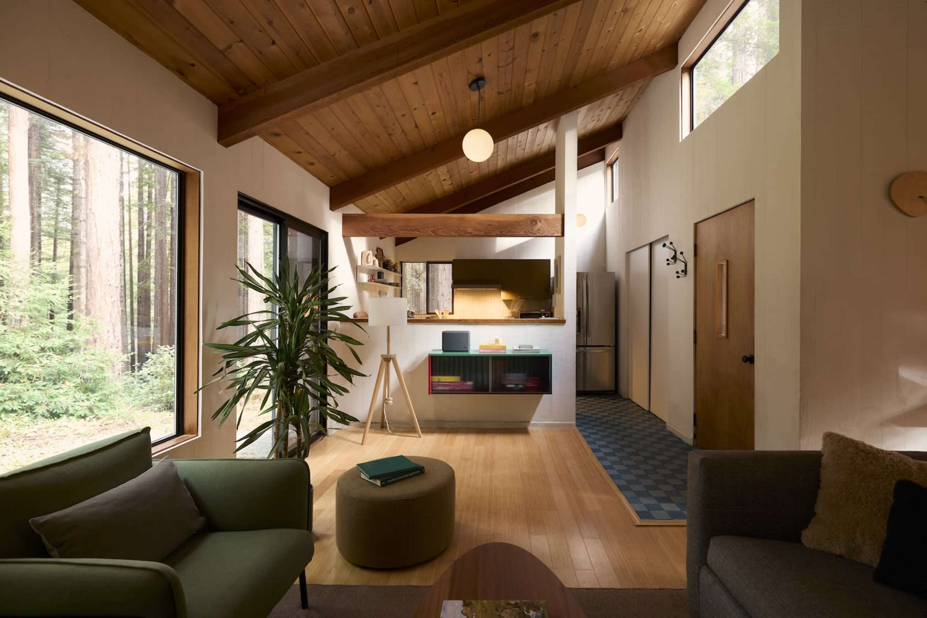 A modern living room with wooden accents, large windows revealing forest views, and a minimalist kitchen area.