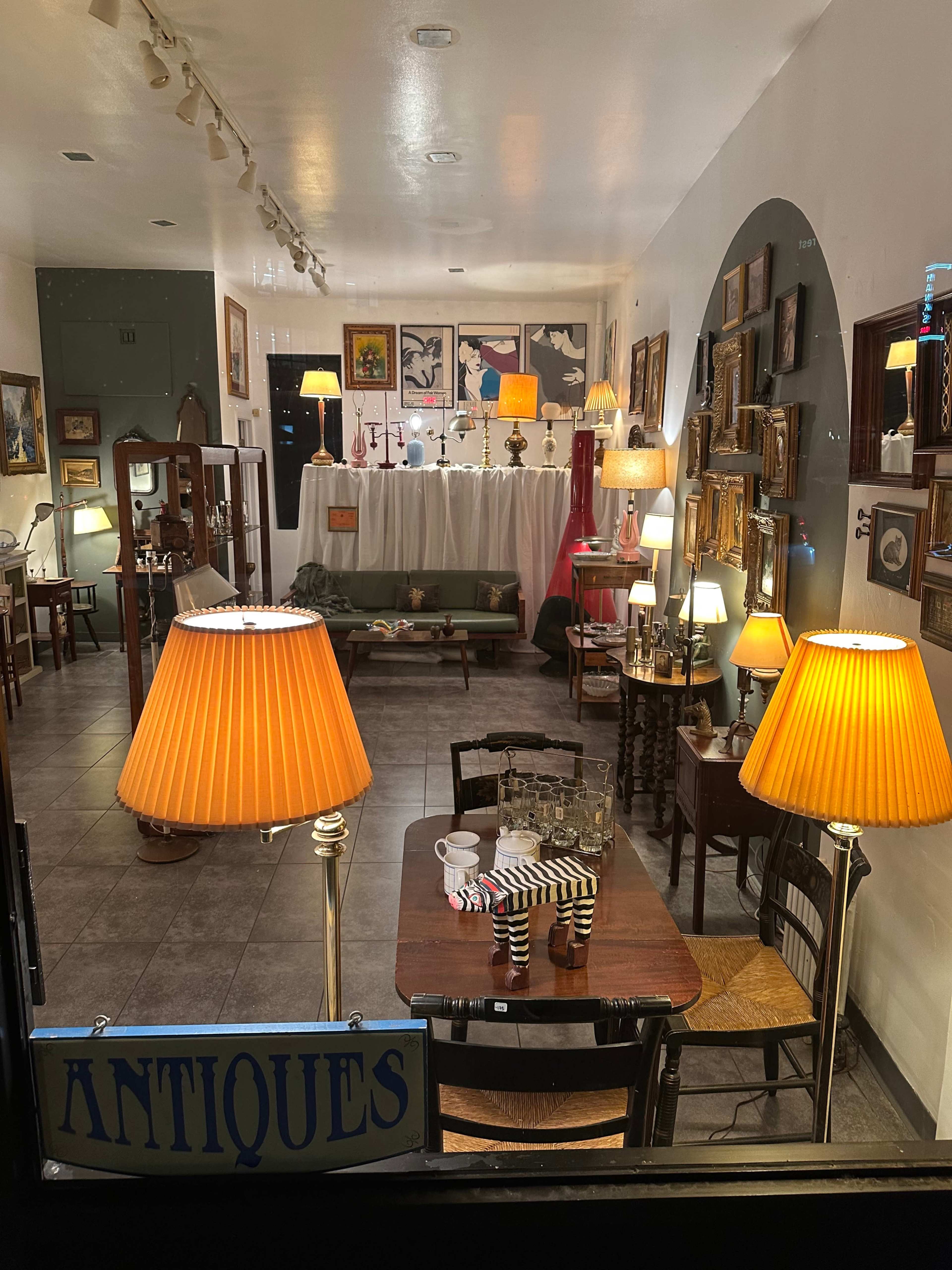 The image shows a cozy antique shop interior with various artworks and lamps illuminating the space, along with a table set with coffee cups and a striped cloth.