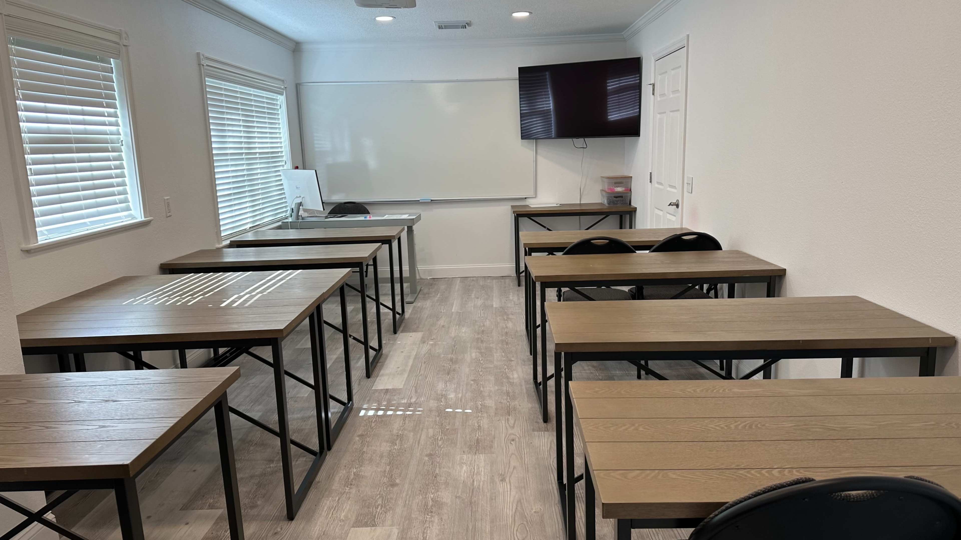 A classroom features several wooden tables arranged in rows with a whiteboard and television mounted on the wall.