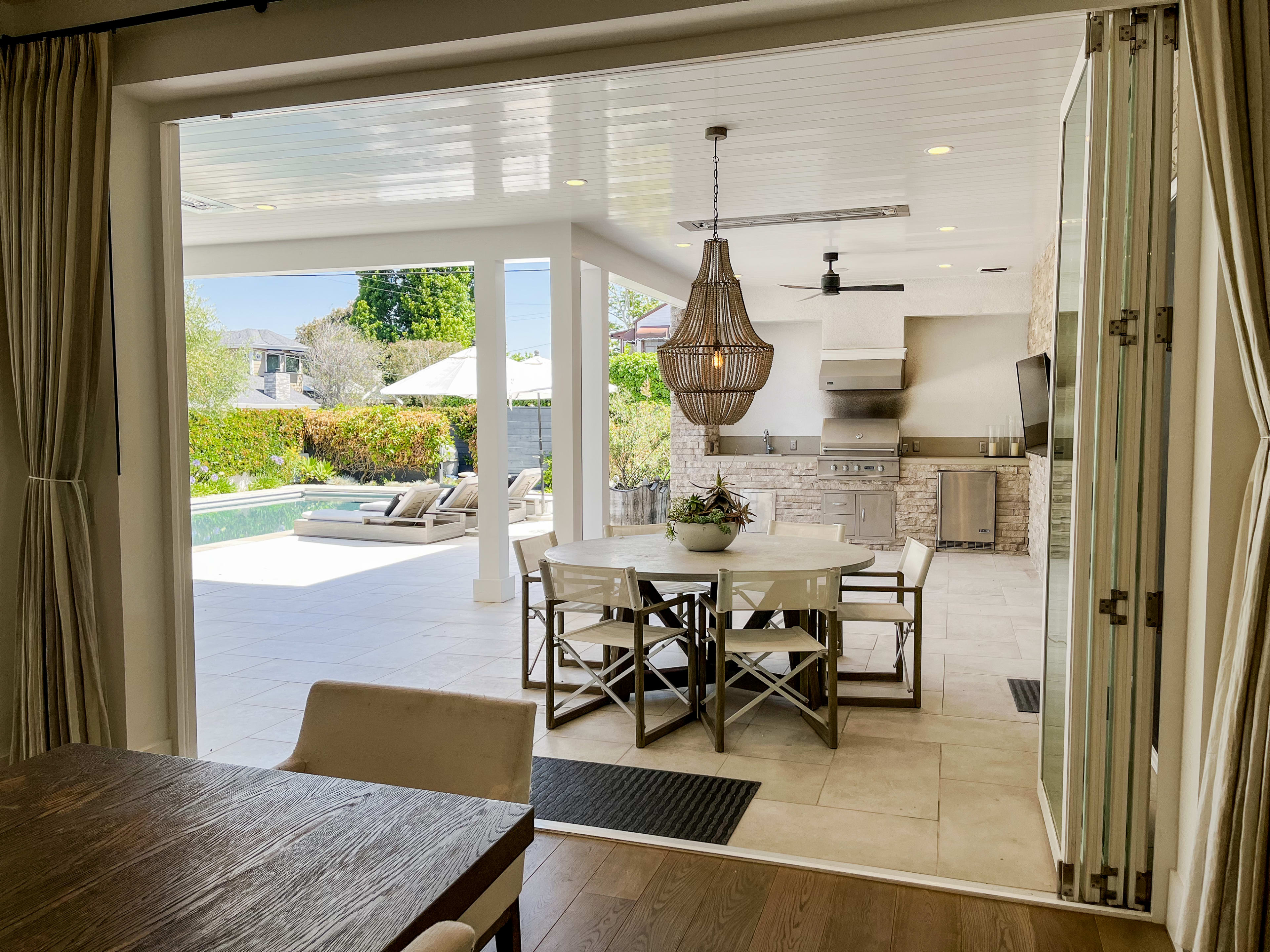 The image shows a spacious indoor dining area with a view of an outdoor kitchen and a pool surrounded by lounge chairs.