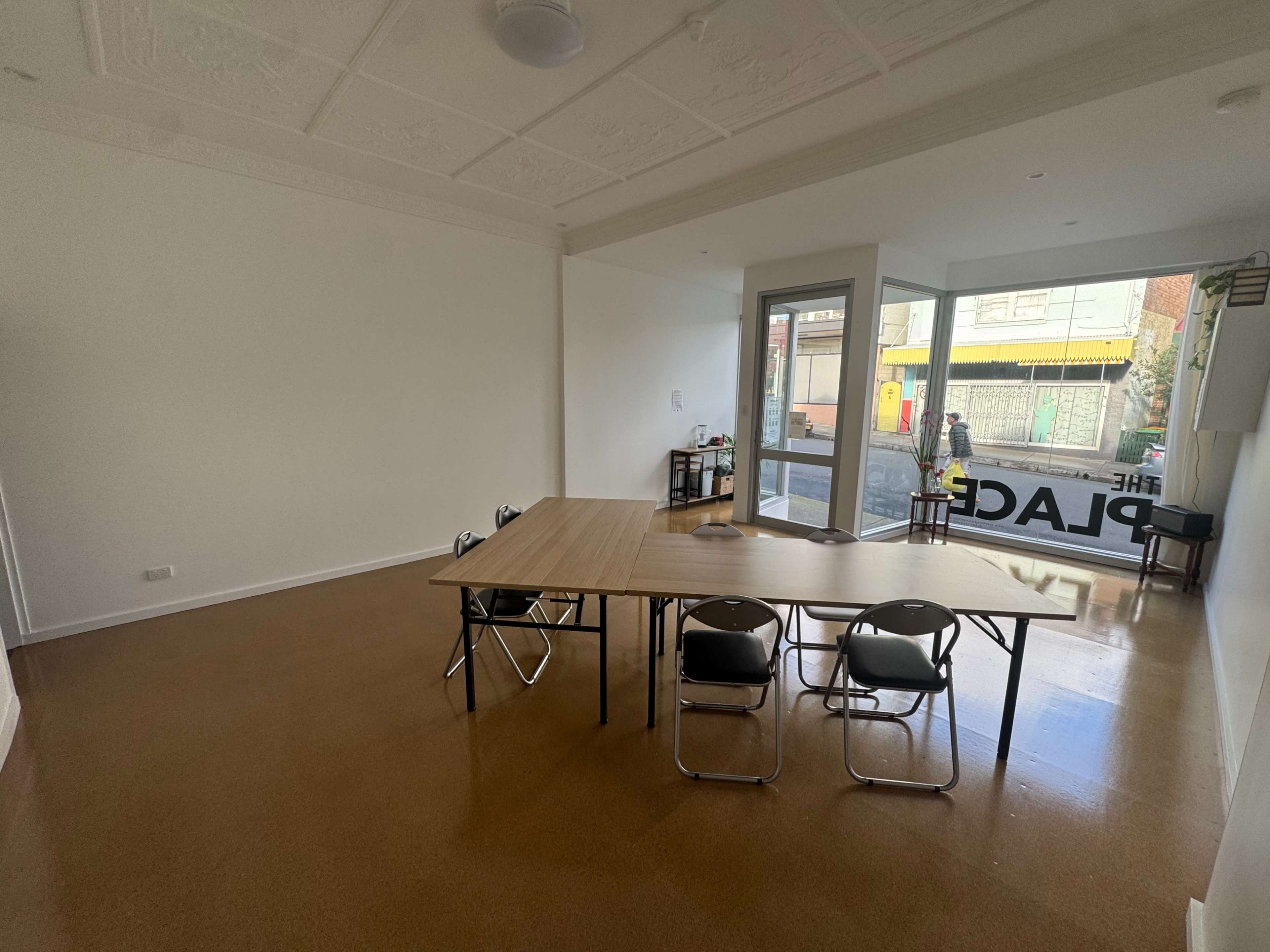 The image shows an empty room with a large table and several chairs set up, facing a glass door that opens to a street view.