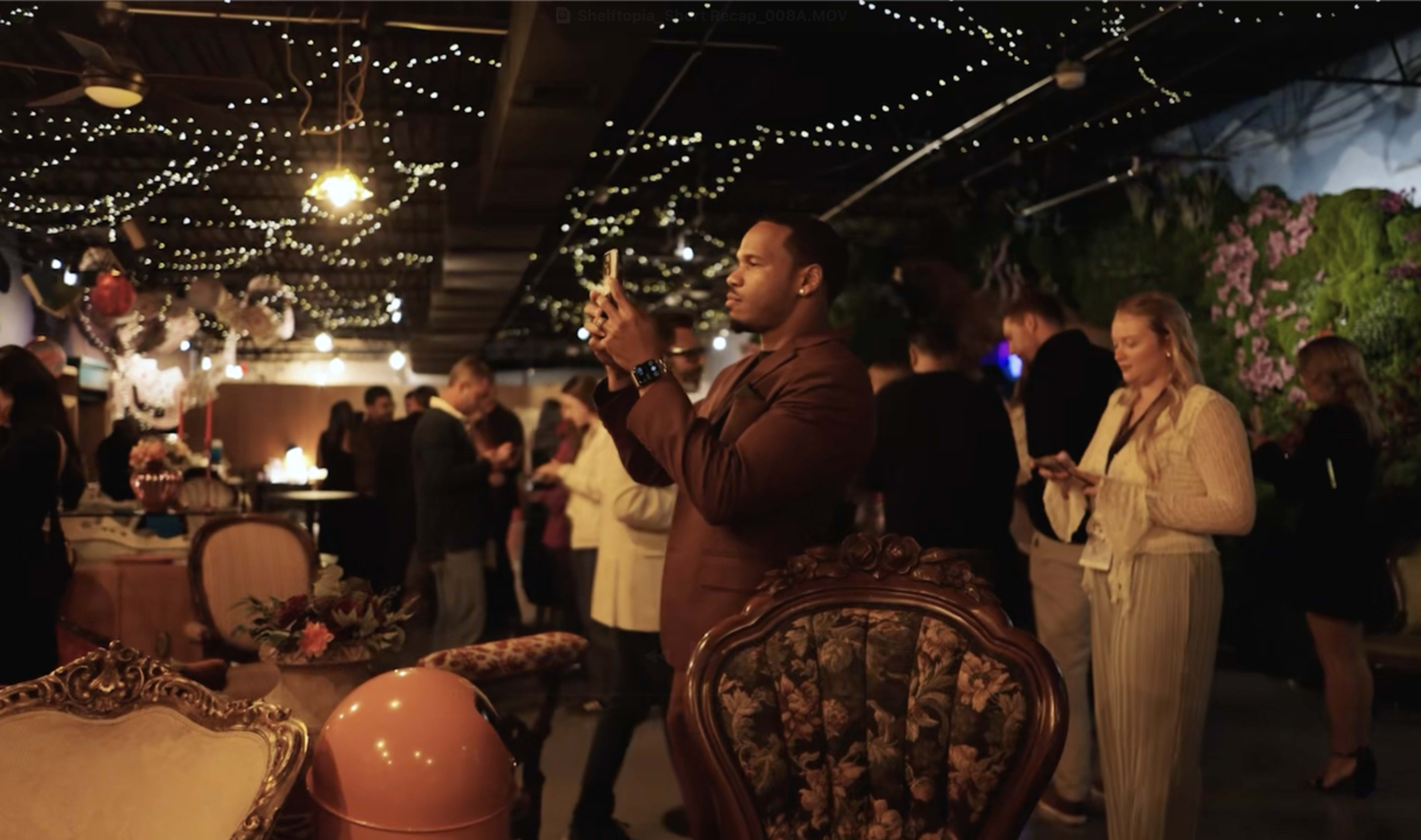 A well-dressed man takes a photo with his phone in a lively venue filled with guests mingling under string lights.